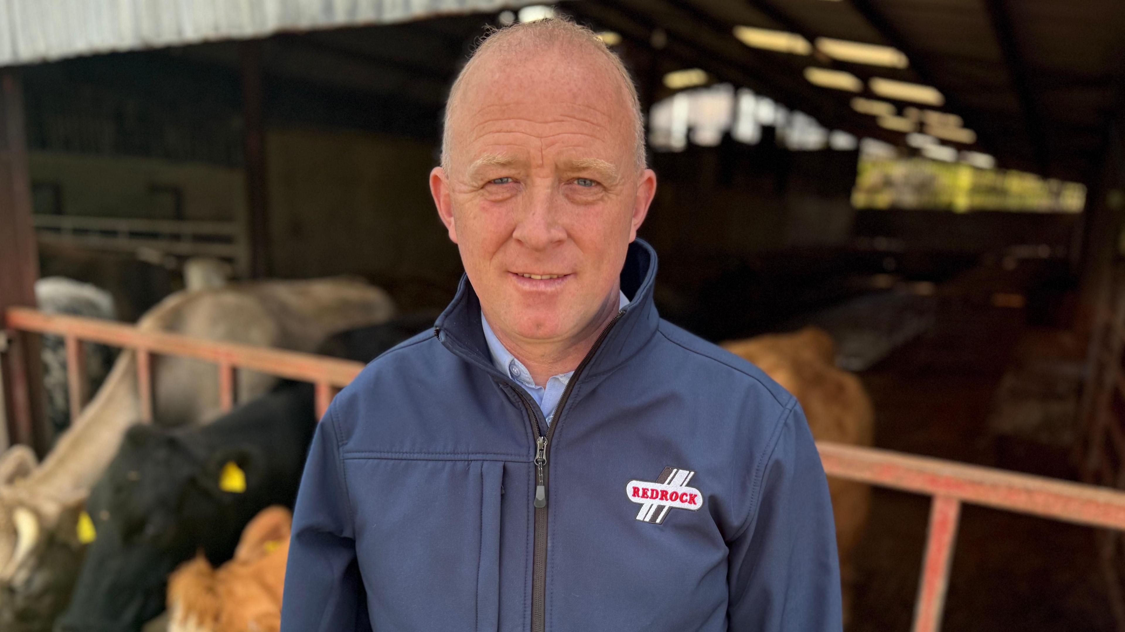 William Sayers, who has thin grey hair, smiles at the camera in front of a cattle shed. He is wearing a navy zip up jacket with 'Redrock' written in red front on his chest. He is also wearing a blue button up shirt underneath. Behind him are three cows, one grey, one black and one ginger, who are blurred.