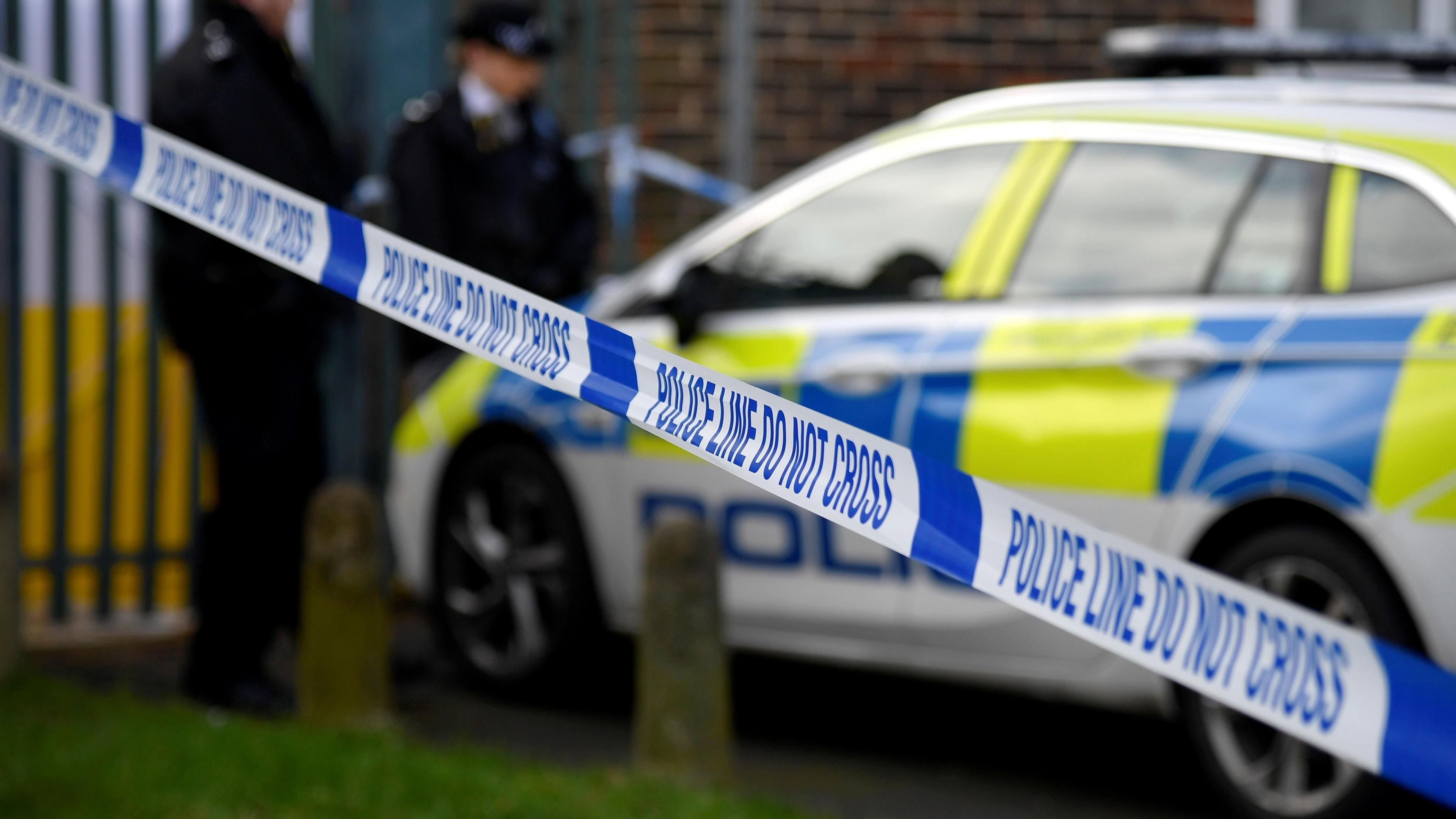 Two police officers, out of focus to the camera, stand by police car behind police tape as they investigate a scene of fatal stabbing, Croydon