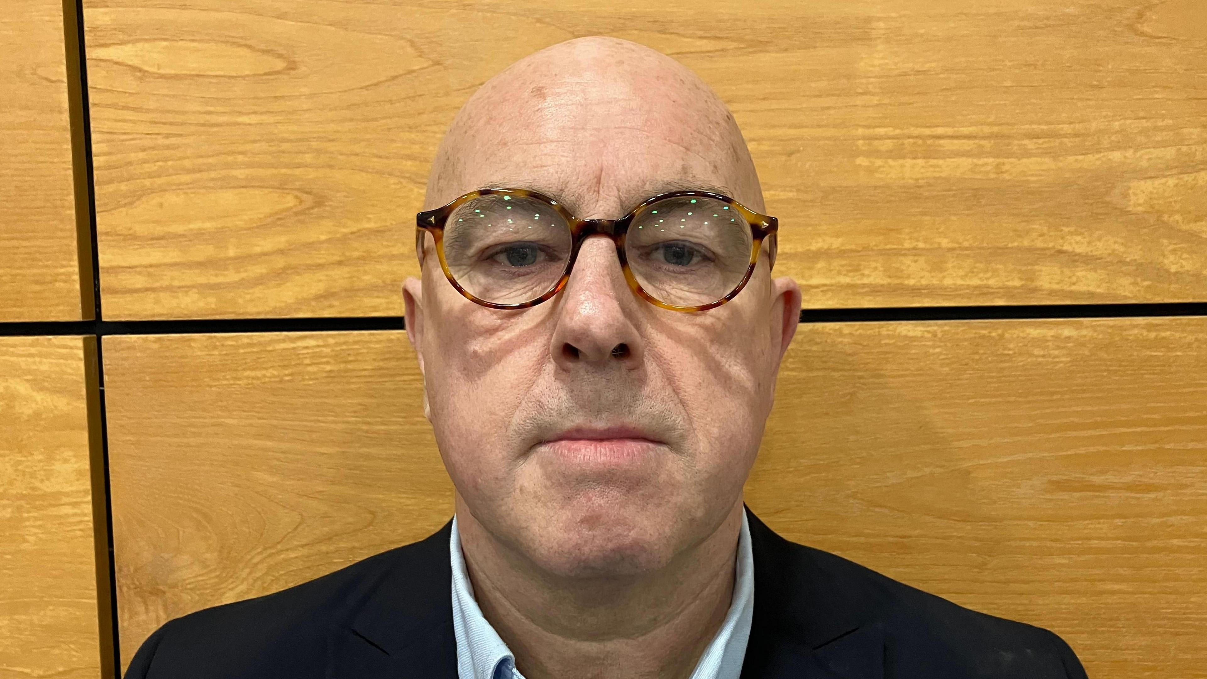 Stephen Maguire is looking straight into the camera. He is bald and has brown rimmed glasses. He is wearing a dark jacket and light blue shirt. There is a wooden wall behind him