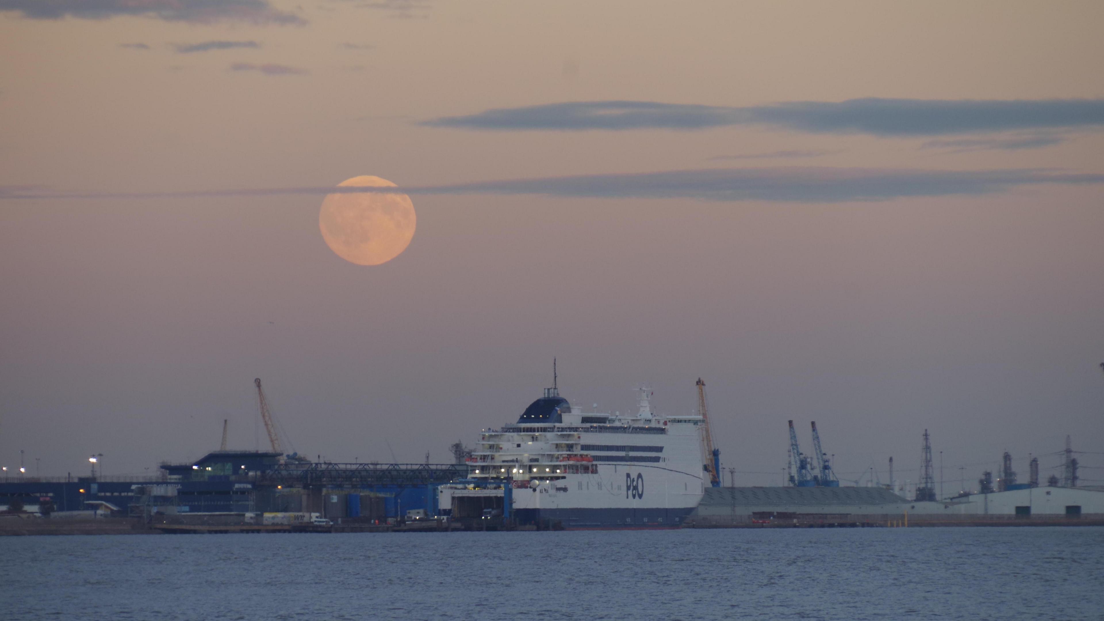A white and blue ferry ship with 'P&O' labelled on the side in blue text. It is docked at an industrial port, with a full Moon in a pastel-coloured sky shining brightly above. There is a blue body of water pictured below the ship.