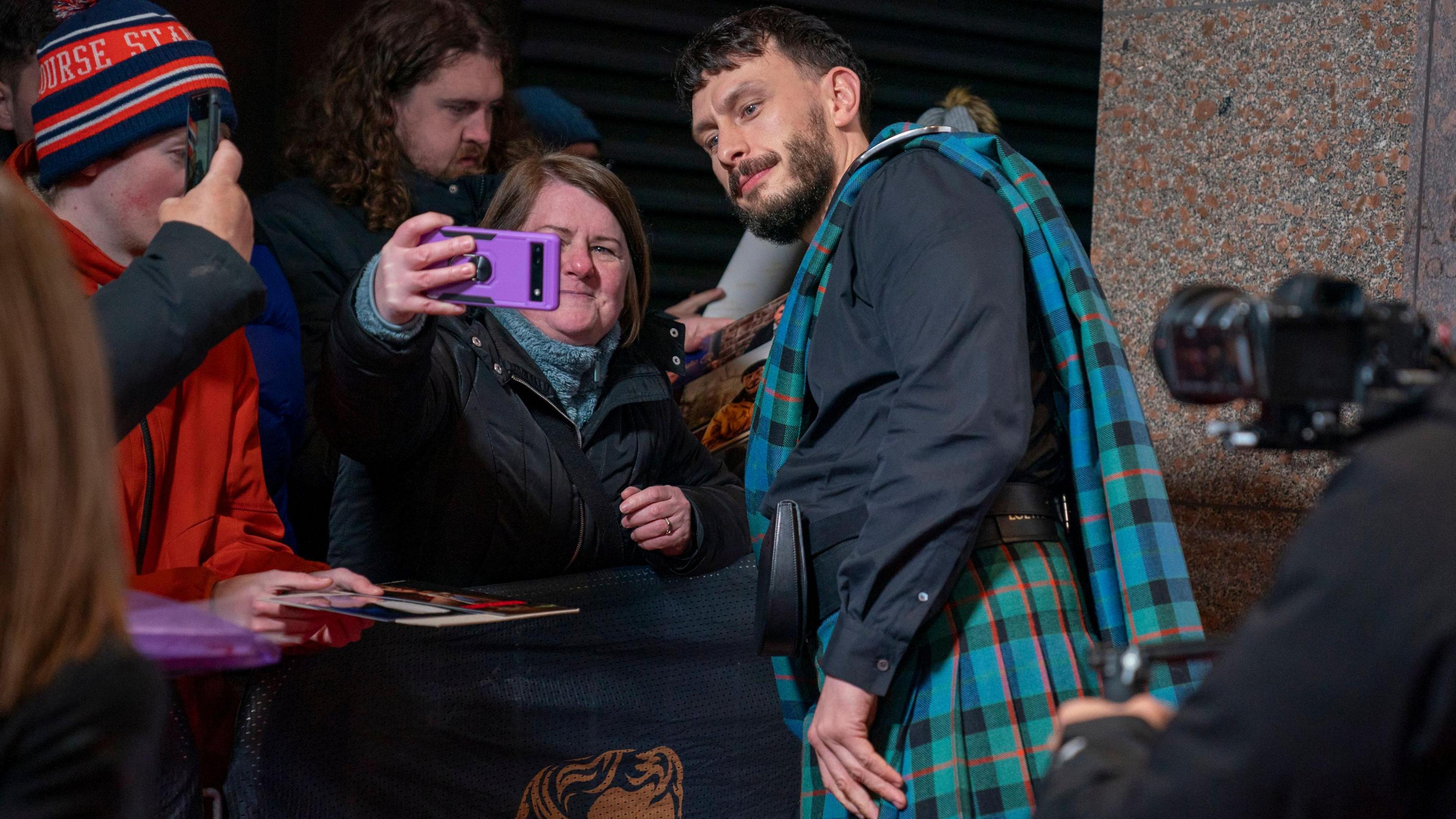 Richard Gadd, wearing a kilt and black shirt, leans towards a barrier to allow a fan to take a selfie on her phone as he arrives at the Bafta Scotland awards. Fans are gathered behind the barrier and a photographer's camera can bee seen in the foreground. 