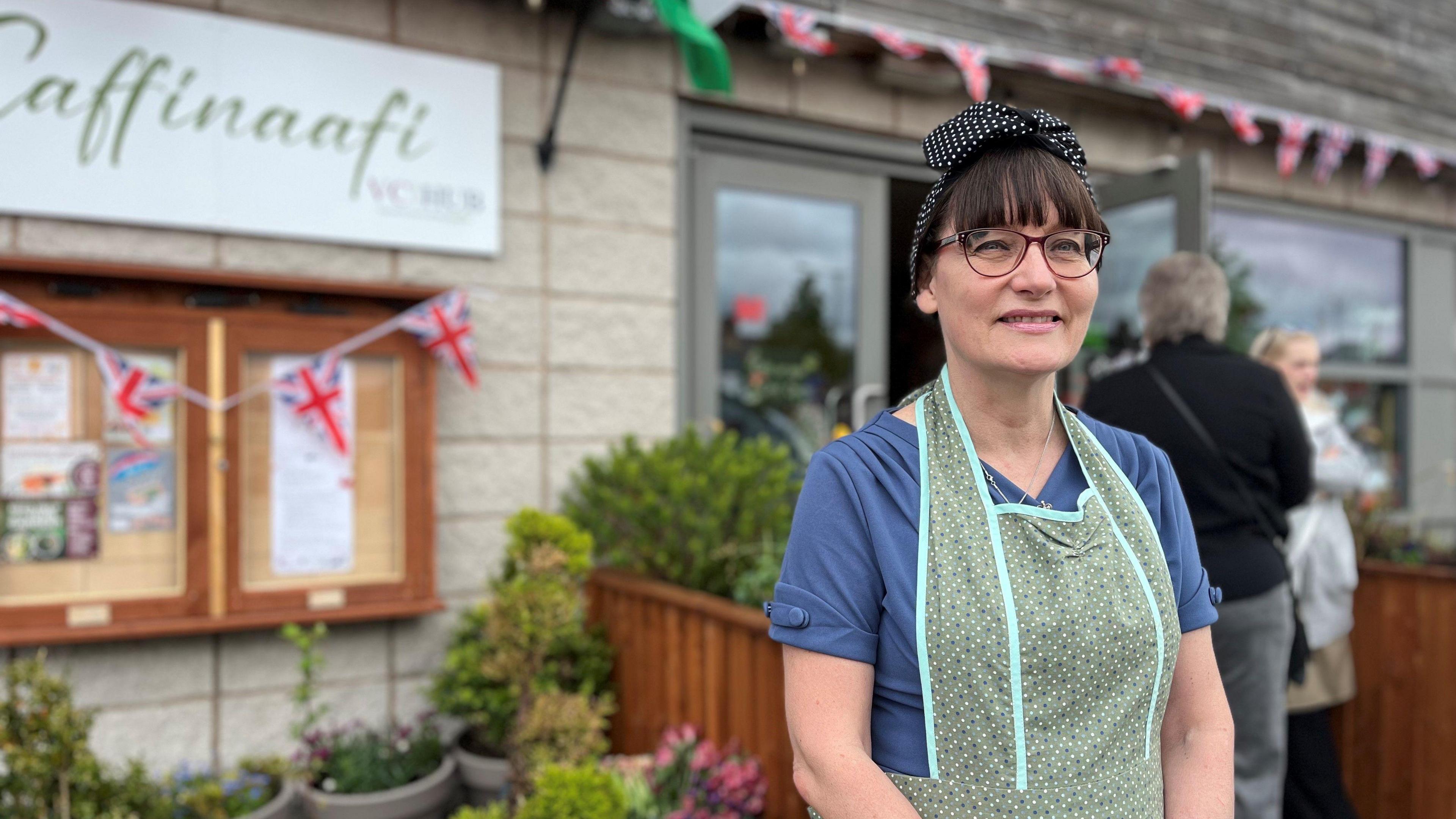 Naomi Horlock is standing in from of the veterans hub, which is draped in union jack bunting. She is wearing 1940 style clothing, with a black and white polka dot bonnet, a green apron and a navy shirt with buttons on the sleeves.
