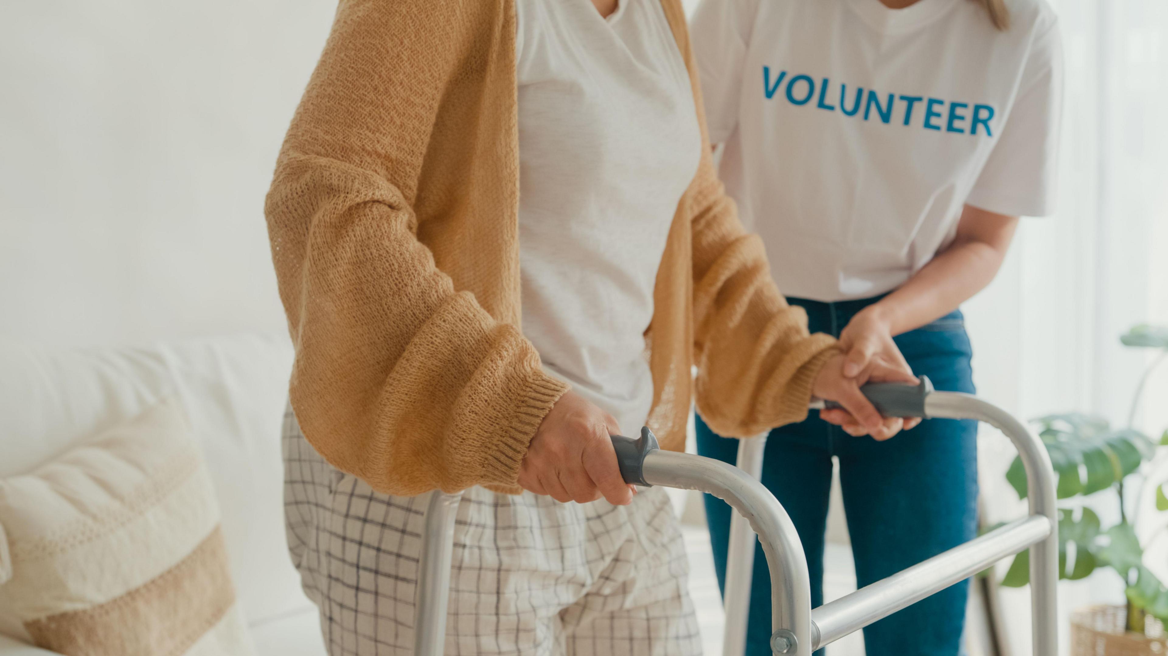Closeup of young female volunteer teach old woman to walk use walker take care hold hand elderly patient in living room at home. The volunteer has "volunteer" written across her t-shirt.