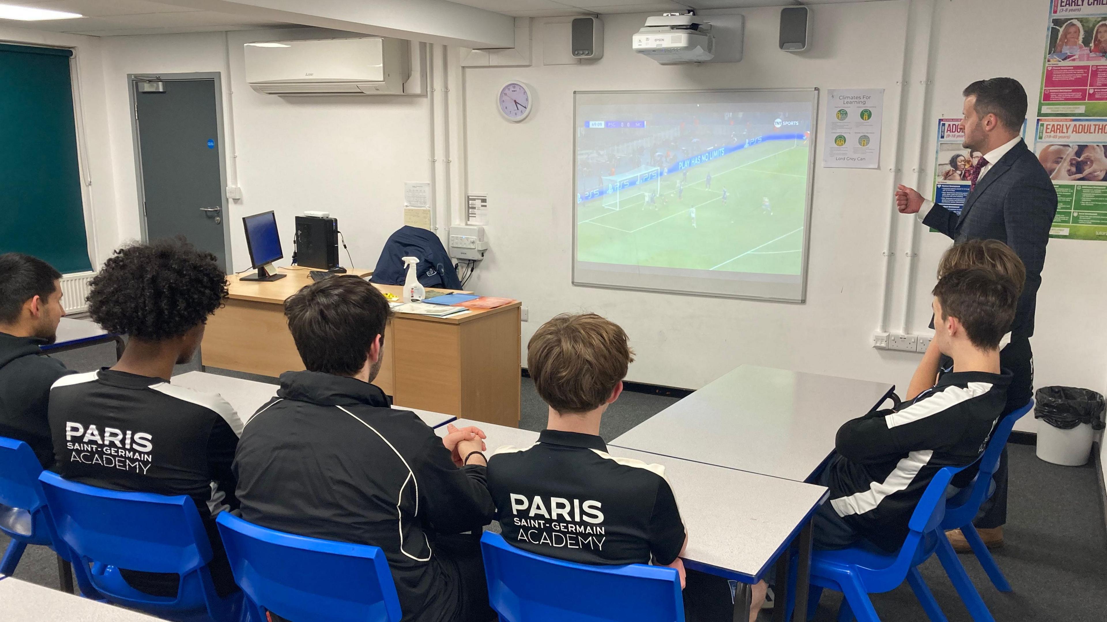 A group of pupils in a classroom watching football on a board while a teacher stands to the side. Two of the boys have "PARIS SAINT-GERMAIN ACADEMY" written on the backs of their tops.