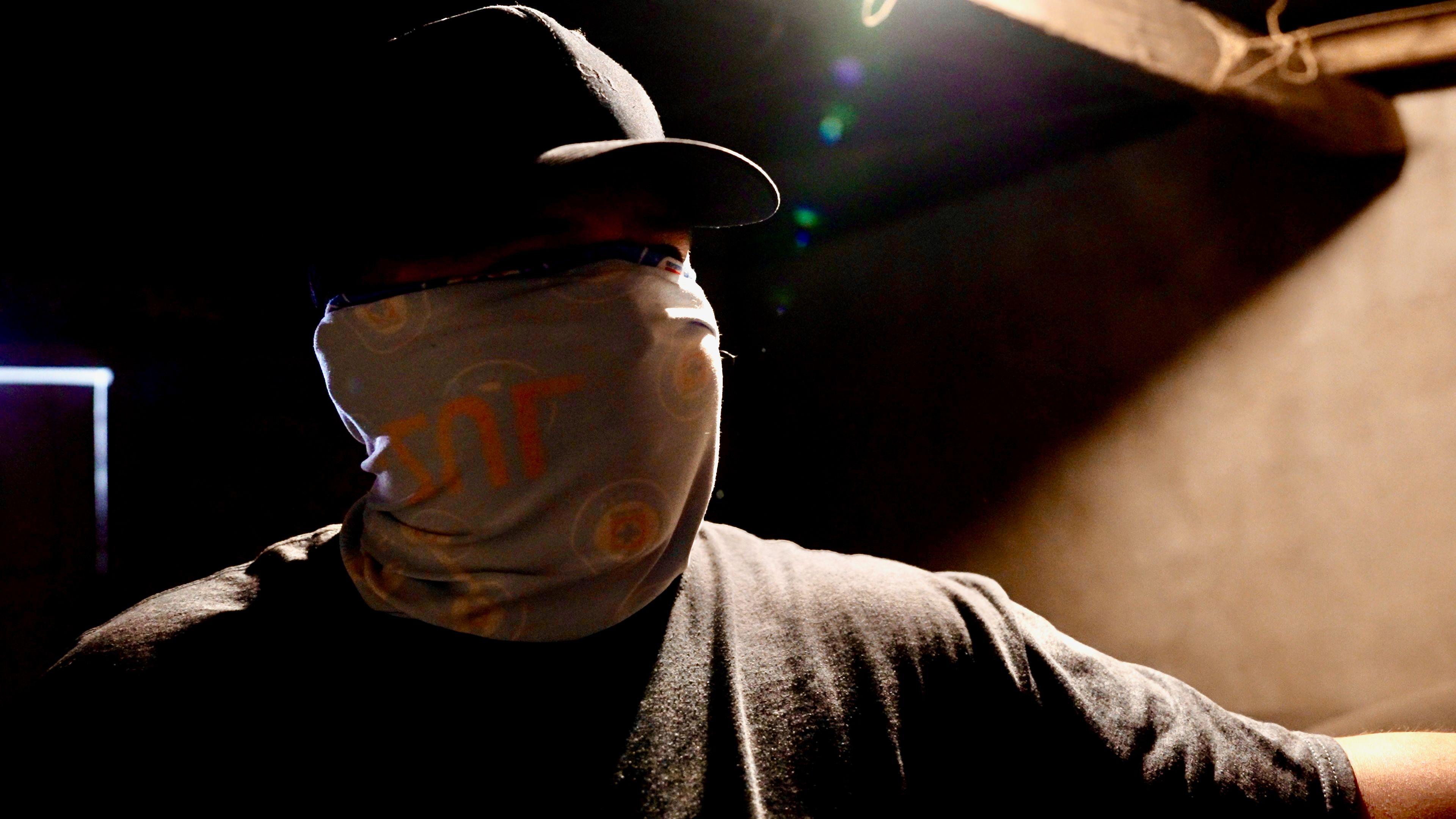 A man wearing a baseball cap and with a bandana covering his face, looking towards the camera as he stands in a bare, starkly lit room, much of which is in deep shadow.