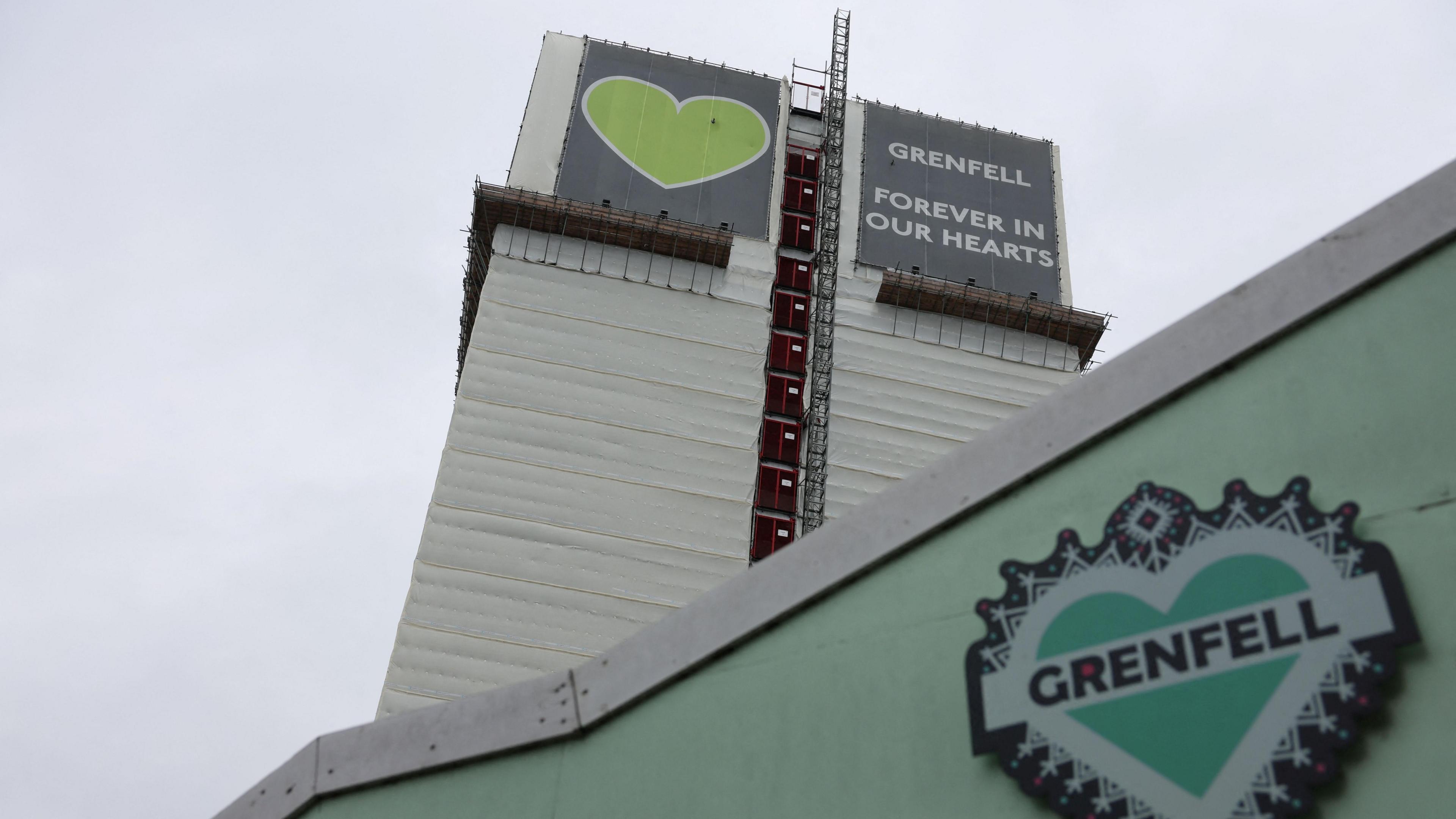 The Greenfell Tower is seen on from a perspective looking up with a green memorial wall seen in the foreground. It features a heart with "Grenfell" written across it. 