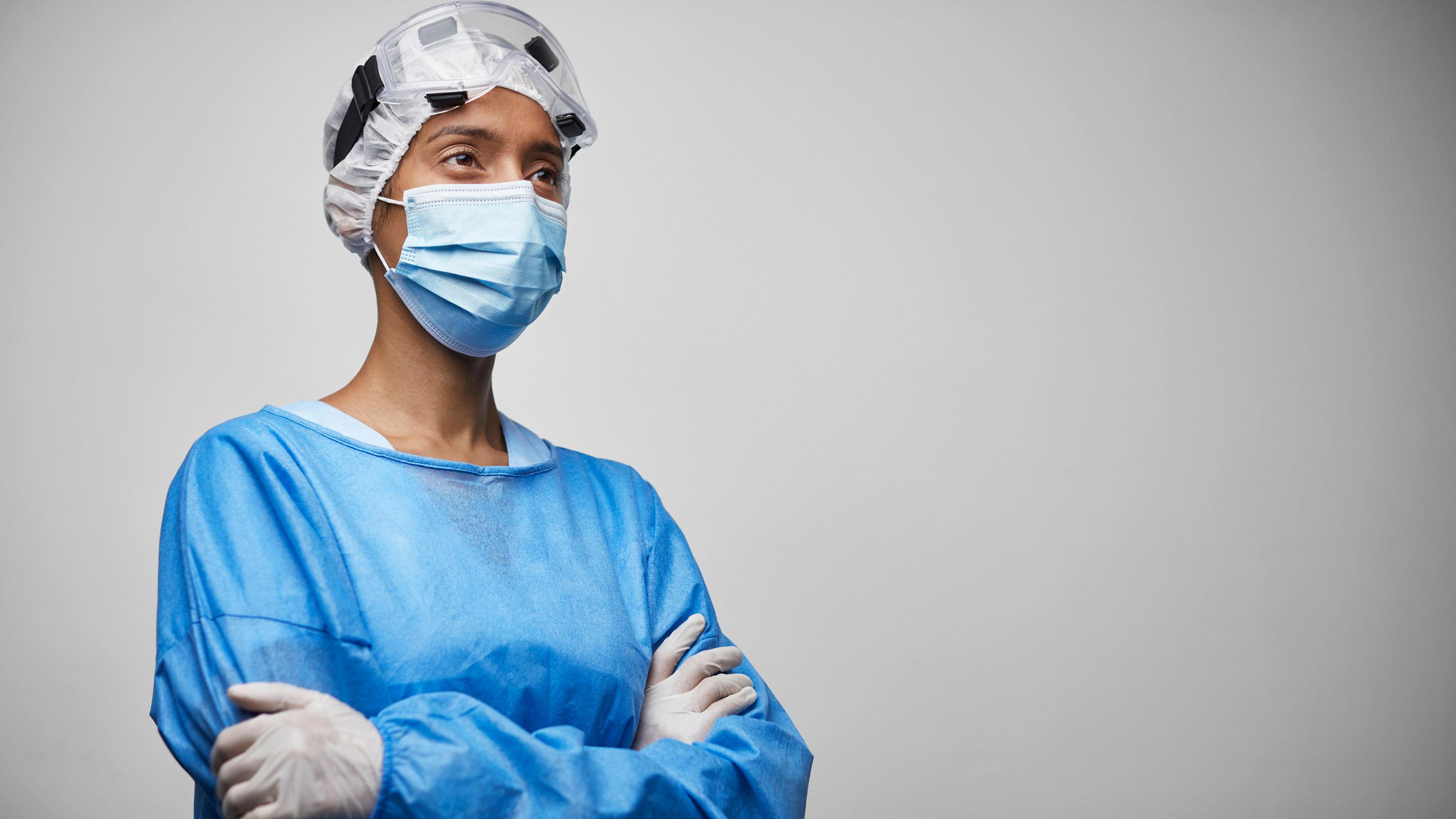Angled view of young female doctor in hospital workwear, bouffant cap, surgical mask and protective eyewear looking away with a serious expression. 