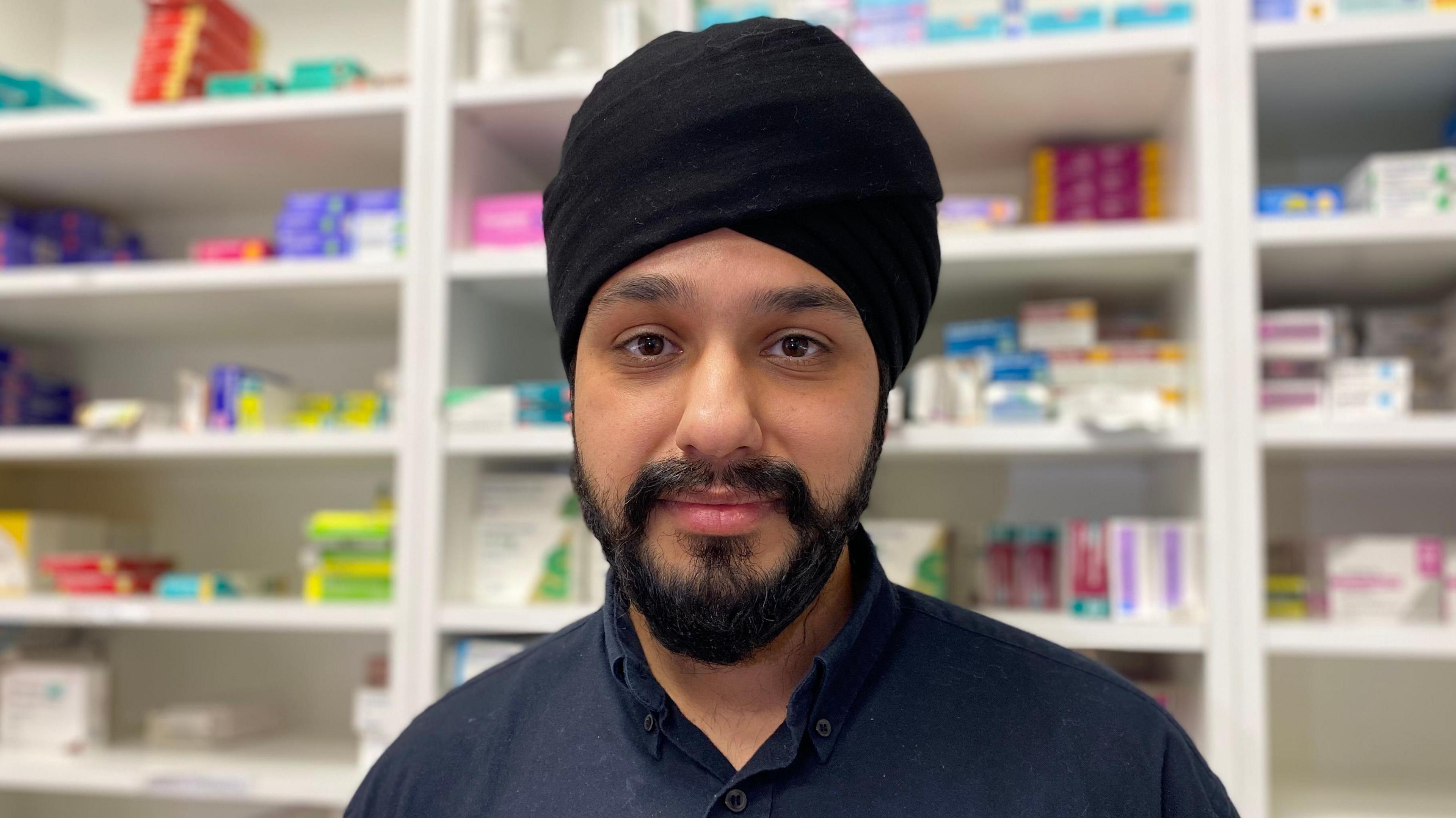 A man with a black turban stood in front of shelves with medicine packets on them.