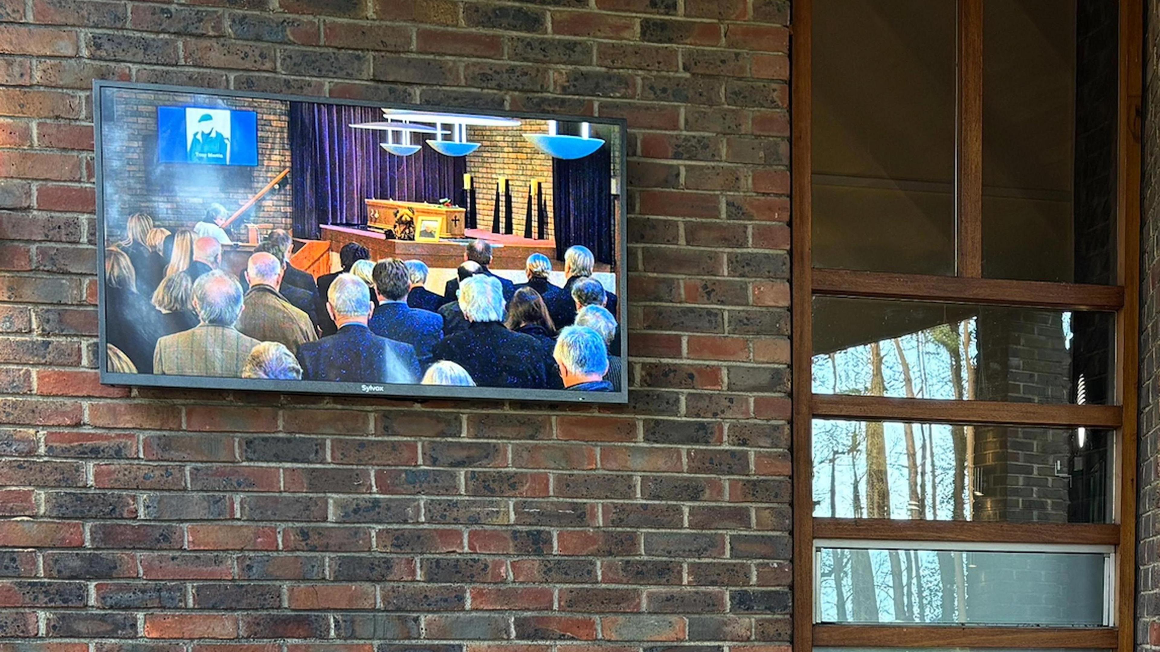 A TV screen fixed to the outside wall of the crematorium shows four rows o people, with the coffin on a plinth and a photo of Tony Martin on a TV screen at the top of the altar.