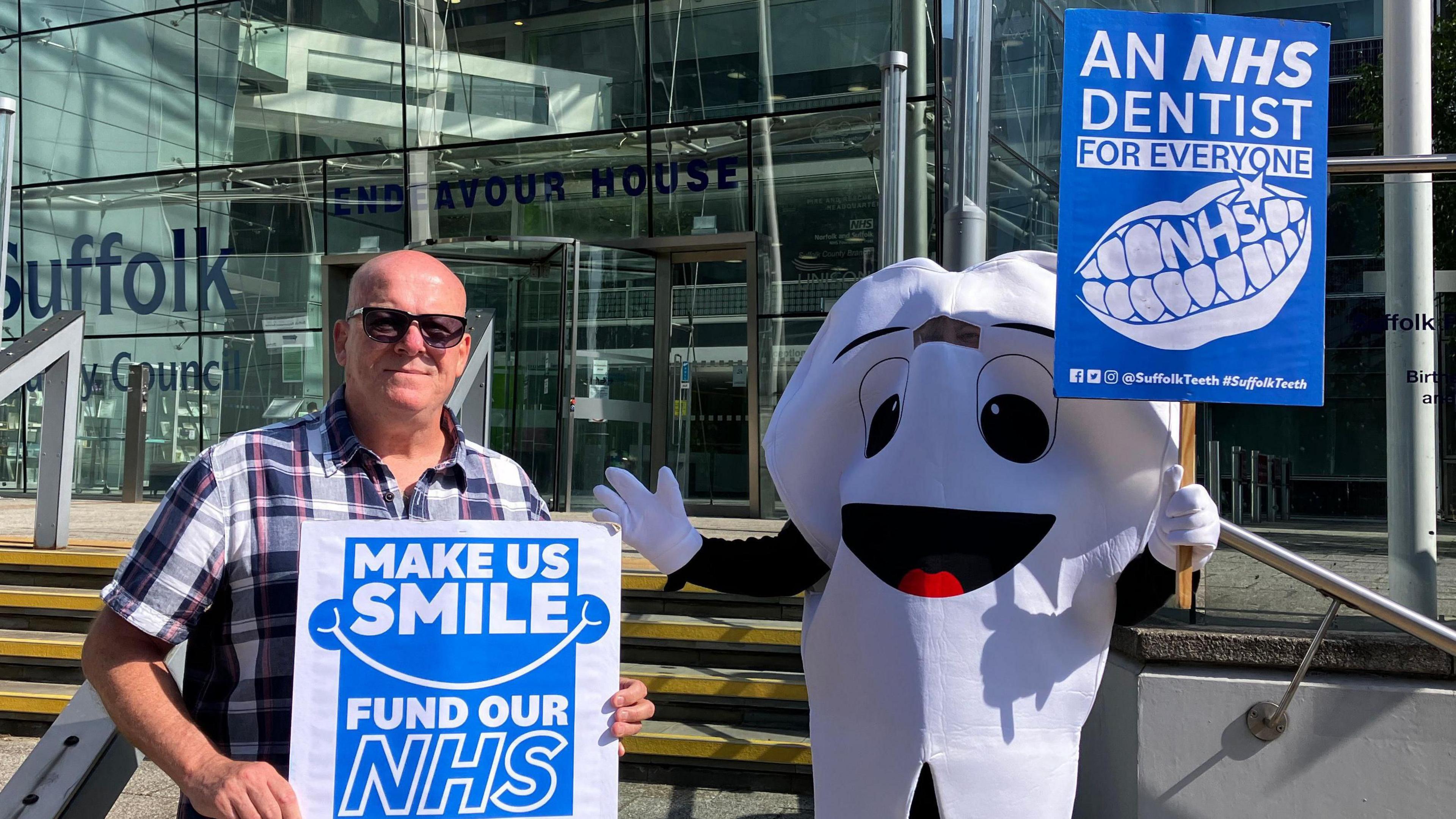 Mark Jones smiles the camera while standing outside Suffolk County Council's headquarters. He is bald and wears sunglasses along with a chequered shirt. He stands next to another person dressed in a tooth outfit. Both are holding up placards calling for more NHS dentists