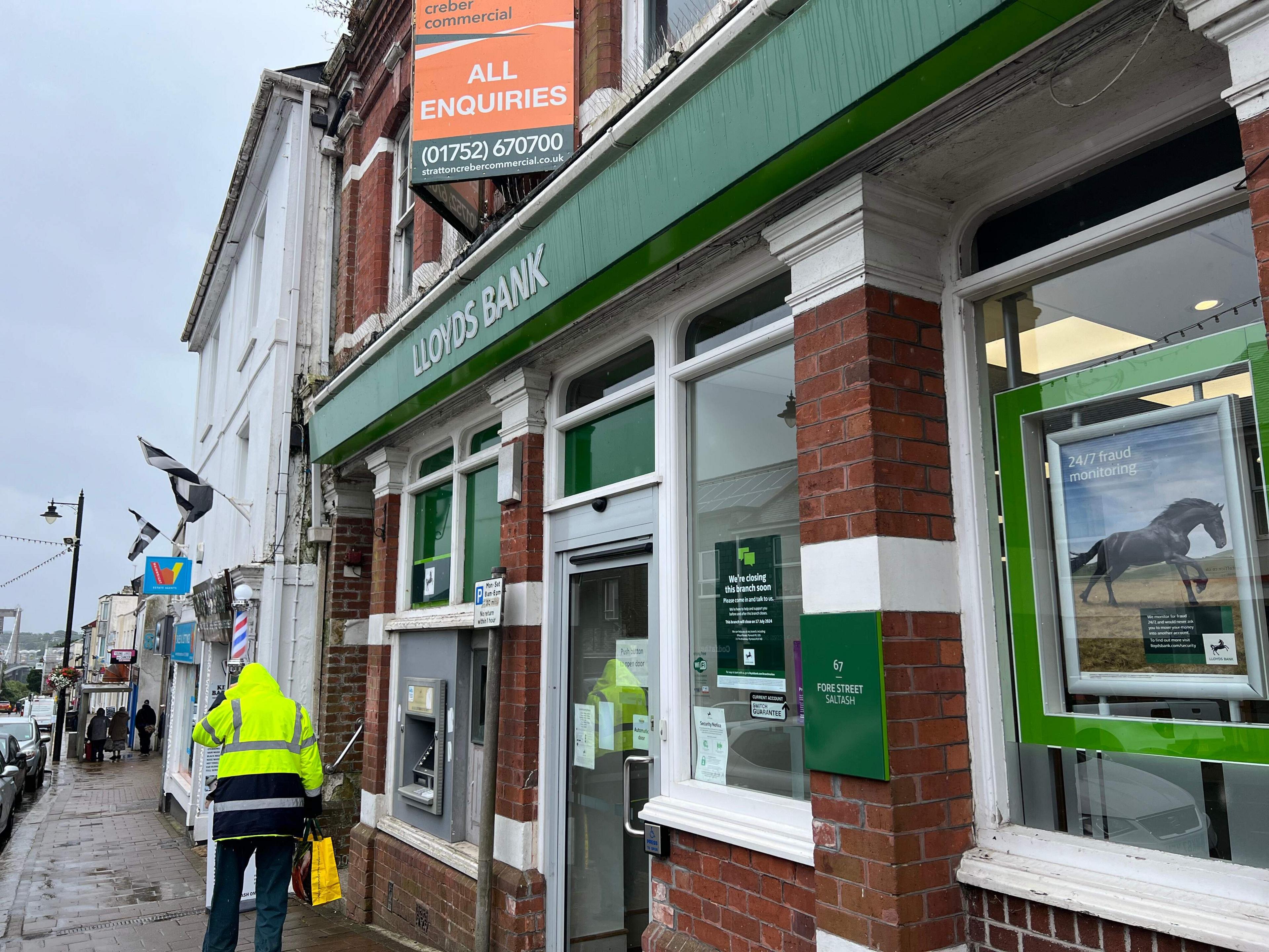 A man in a high vis yellow rain jacket walks past the bank building on Saltash high stree. An all enquiries sale board is visible above the bank's branded sign.