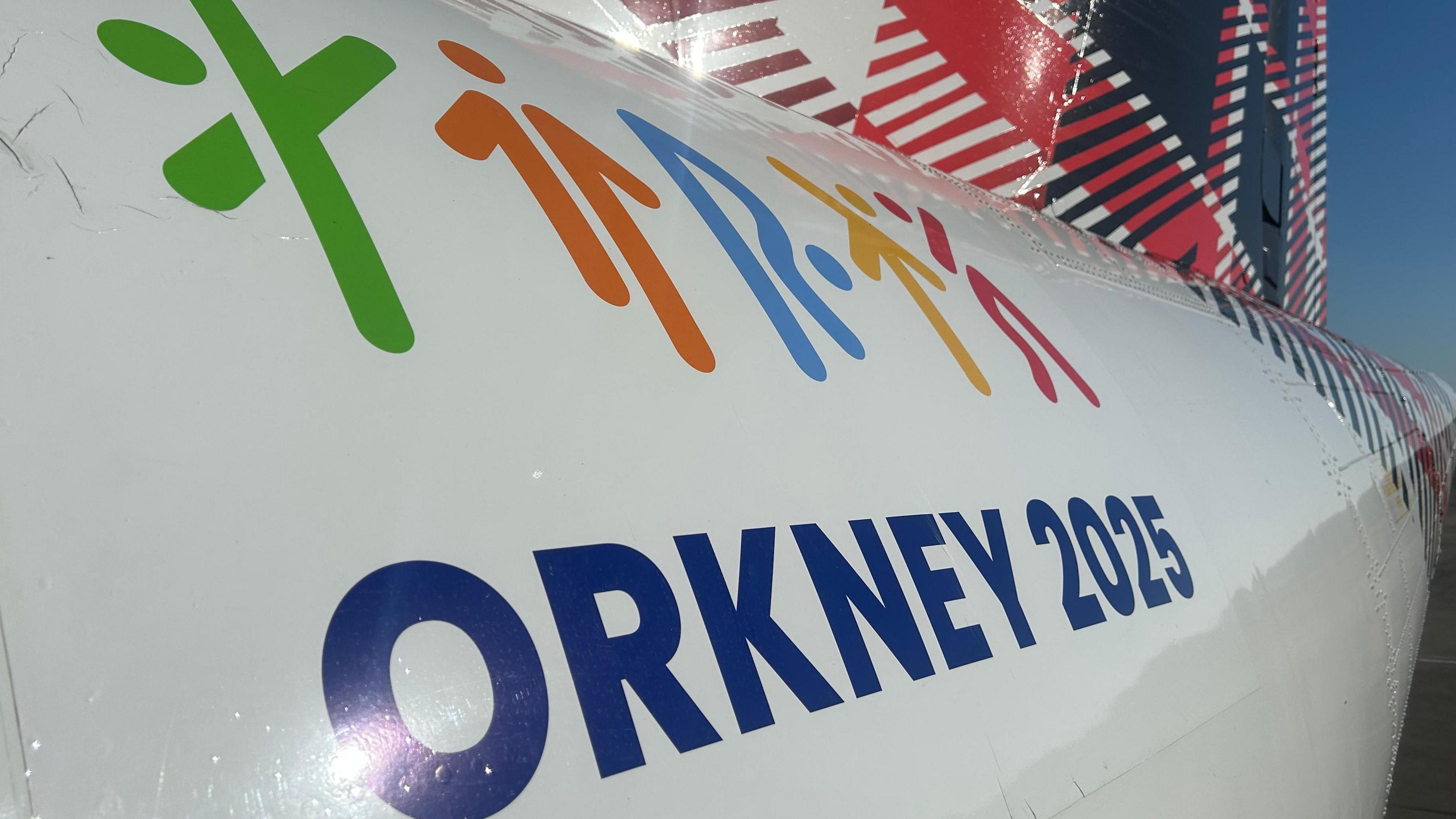 Livery on the side of an airplane which reads Orkney 2025 and has five stickmen in different colours taking different poses. There is also a tartan pattern on the tail.