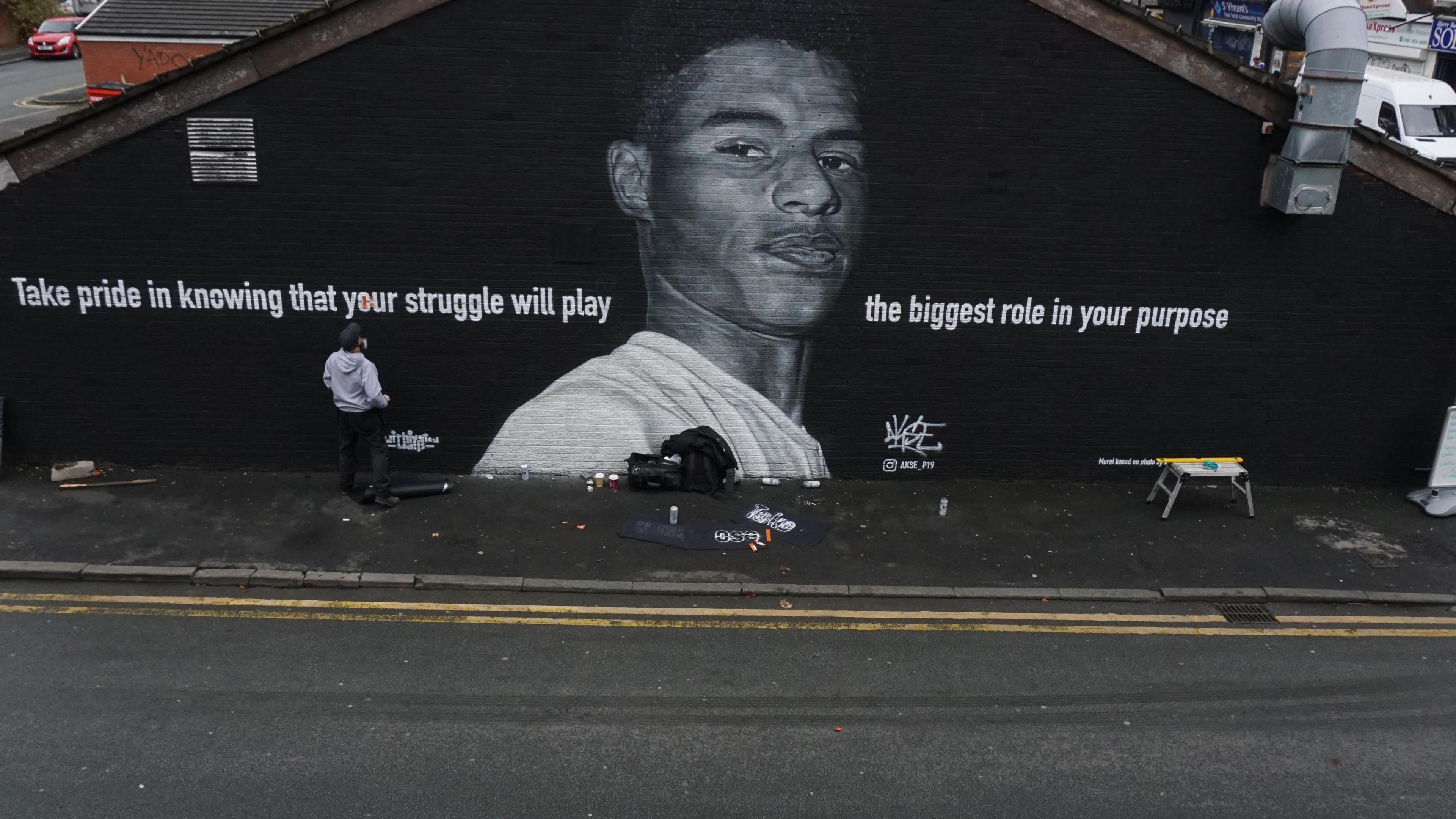 A mural of Marcus Rashford where his face is painted in black and white on top of a black brick wall, with the quote 'Take pride in knowing that your struggle will play the biggest role in your purpose'