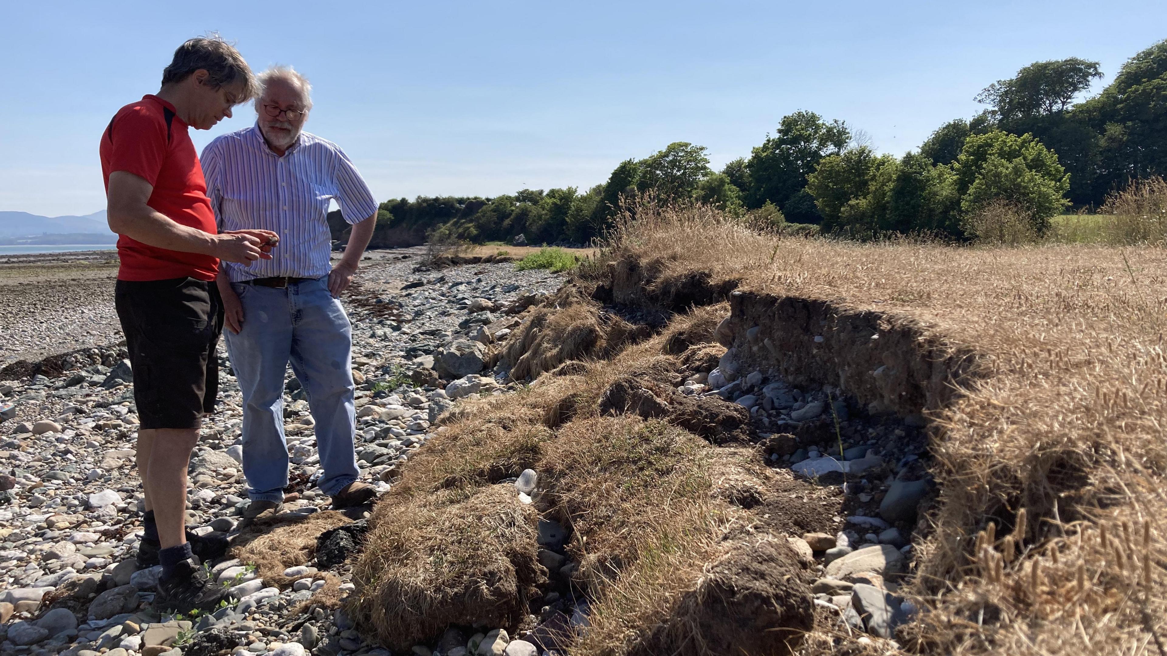 Two men examine the coastal erosion of the land as it meets the beach