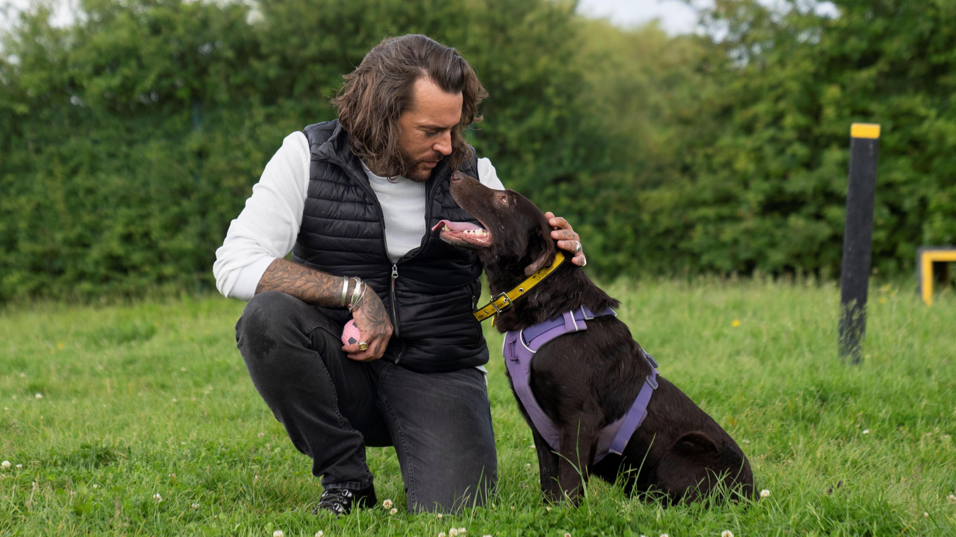 Pete Wicks, wearing a black gilet, white long-sleeved shirt and dark trousers, crouches down to stroke a chocolate-brown Labrador who gazes up at him with its tongue lolling out