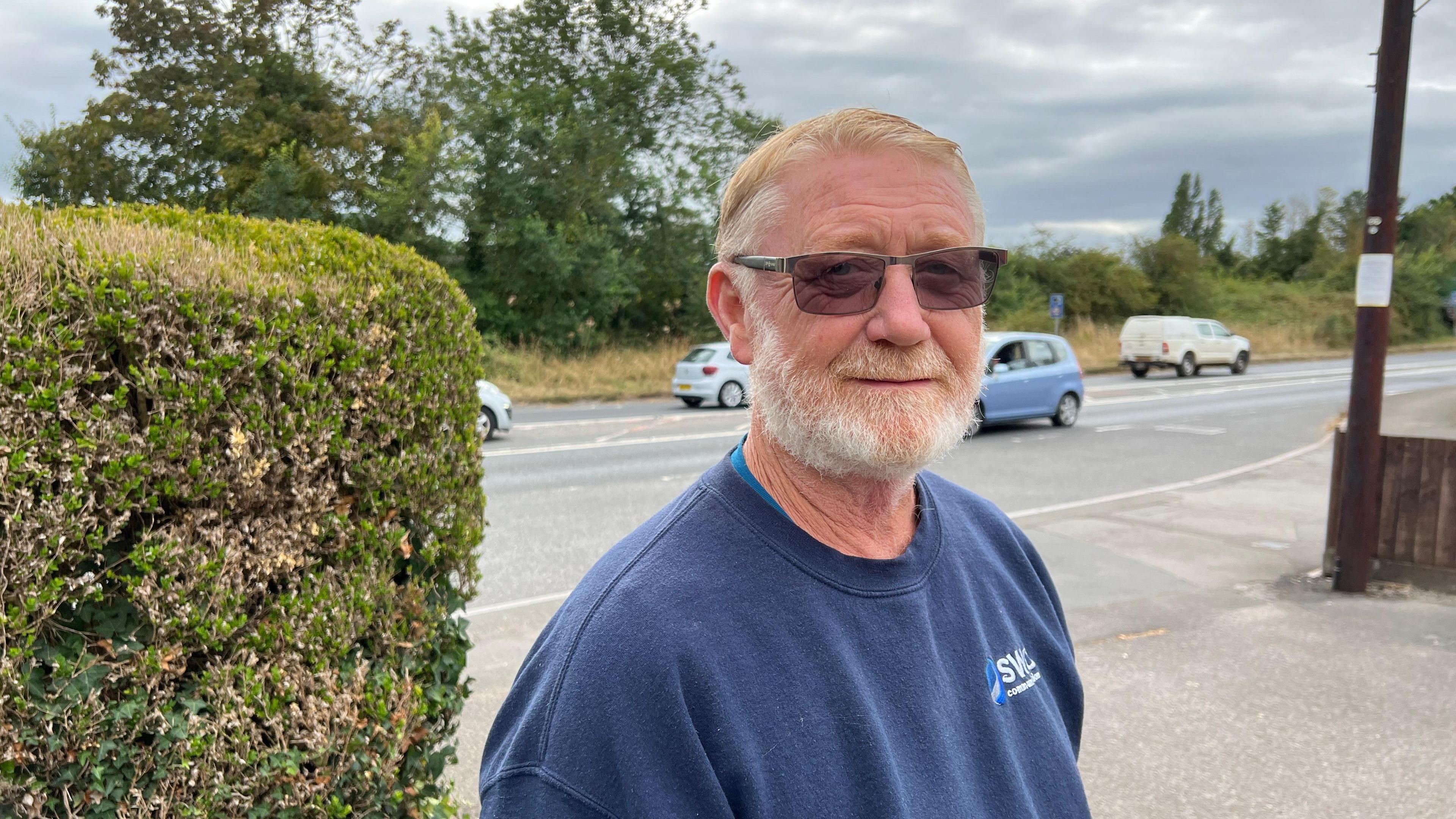 Man looking at camera with A358 road in the background. He has a beard, dark glasses and is wearing a light blue sweatshirt