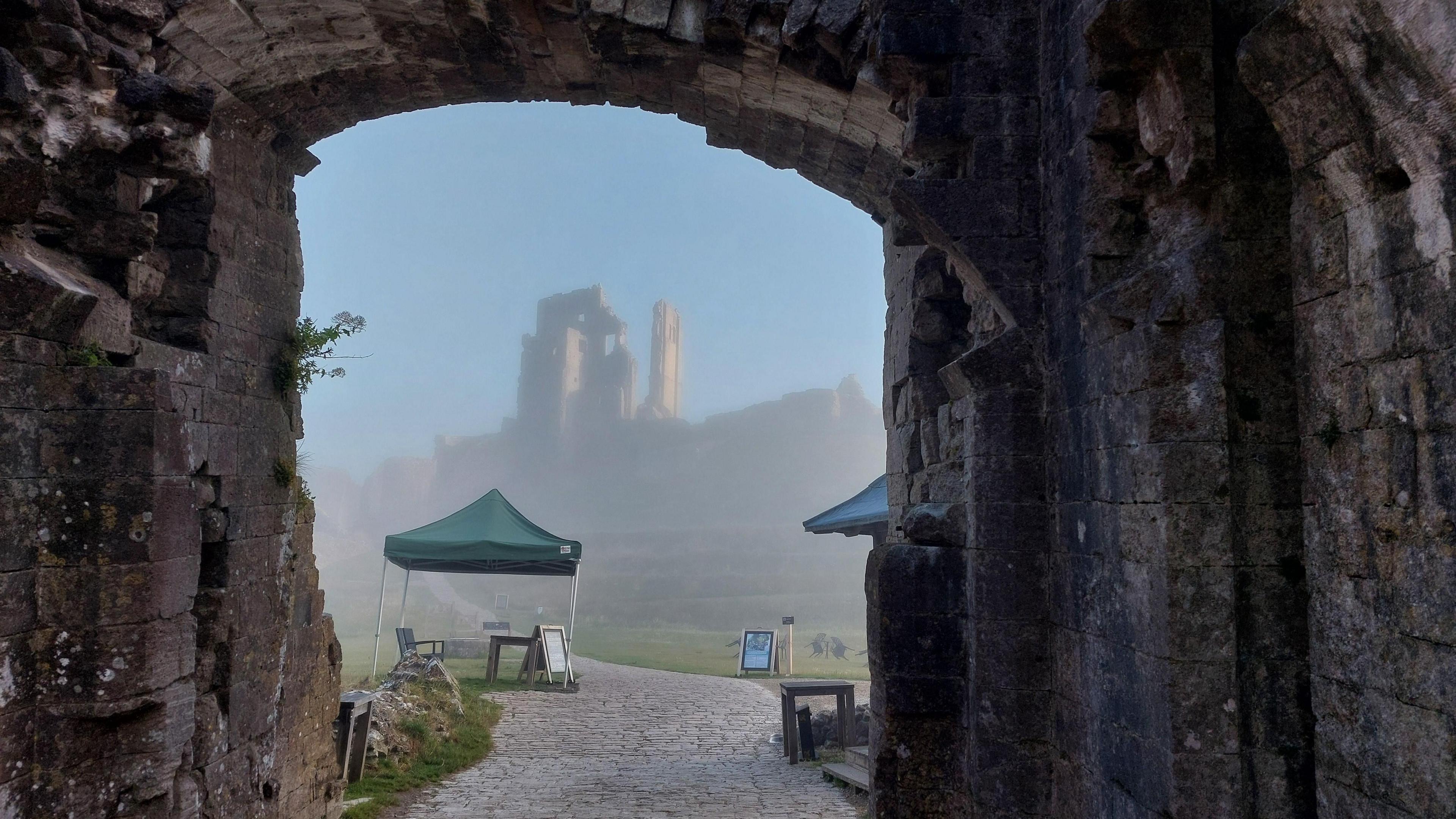 Taken through a stone gateway framing the ruins of Corfe Castle appearing from the early morning fog. In front of it stands an gazebo with a table and chairs by the pathway leading towards the castle.