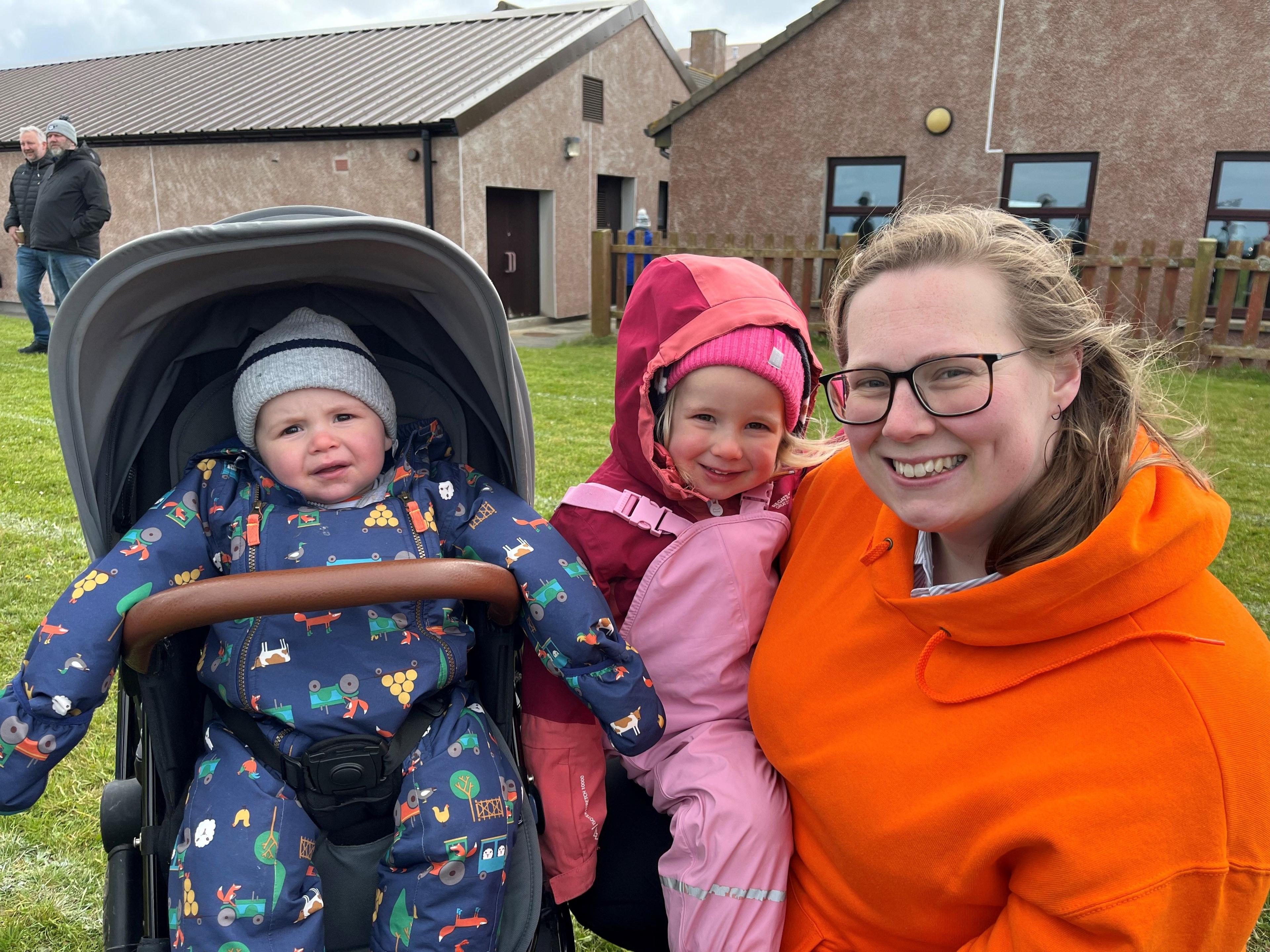 A woman in an orange hoodie kneeling next and holding a toddler with a pink hat on. A boy in a boilersuit with a hat on in a pram.