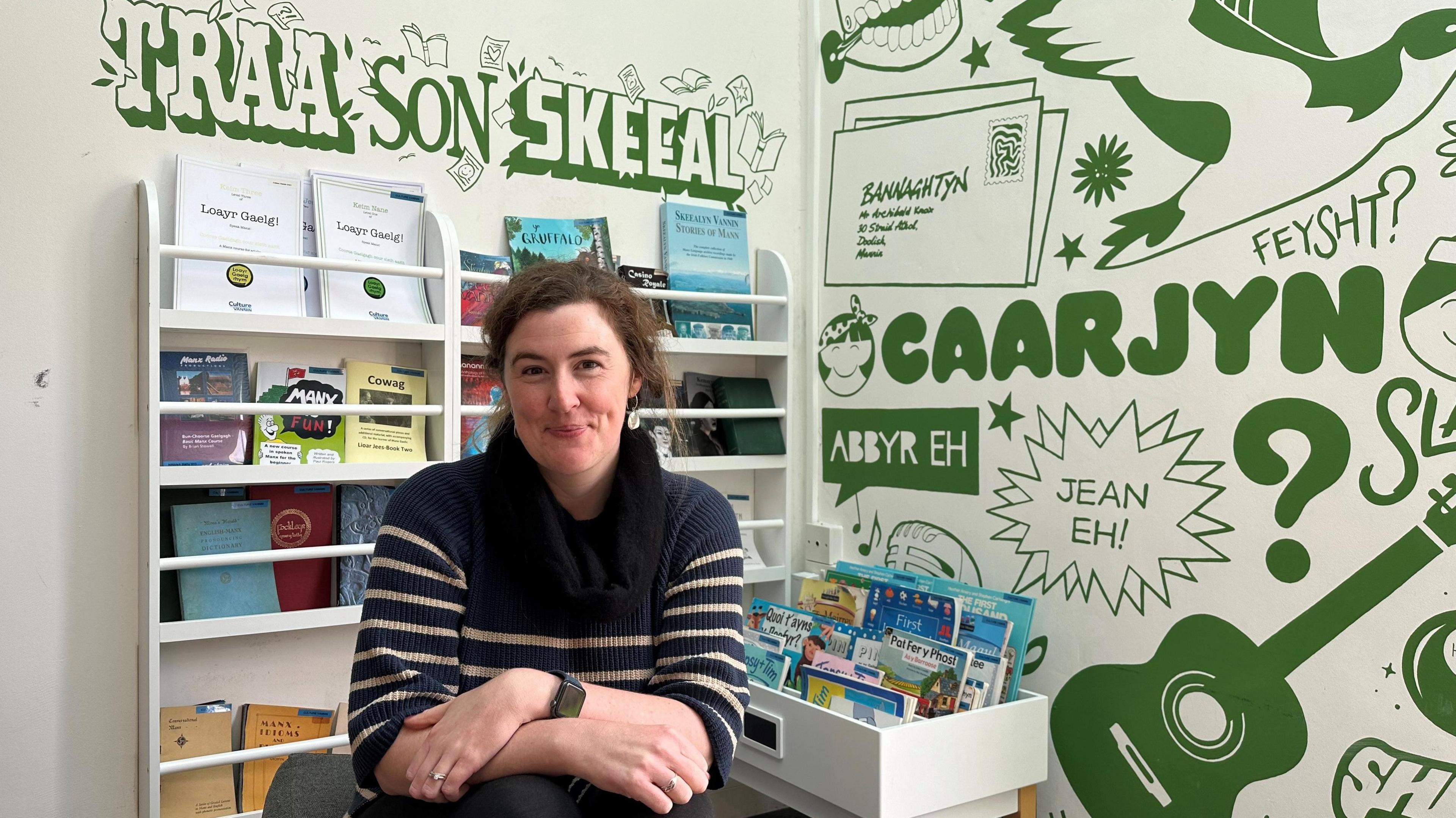 A woman sitting in a classroom with textbooks on a stand behind her, and a green mural with Manx language phrases written on the wall. 
She has brown wavy hair, and is wearing a blue sweater with white stripes. 