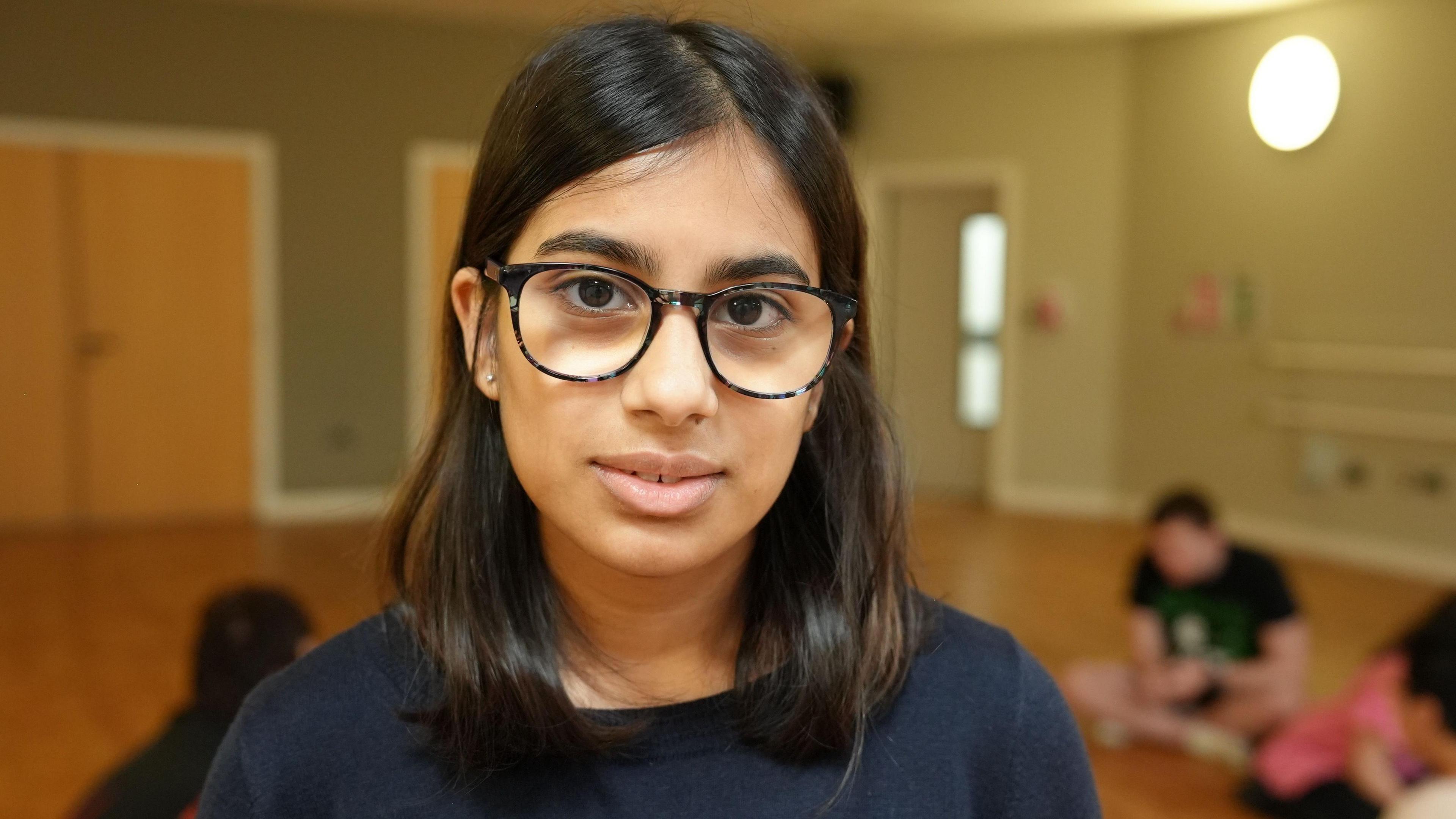 A girl with dark hair and glasses smiles at the camera, while students sit behind her in a dance studio on a light wooden floor. The background is slightly blurred but some wooden doors are visible, and a bright light on the wall.