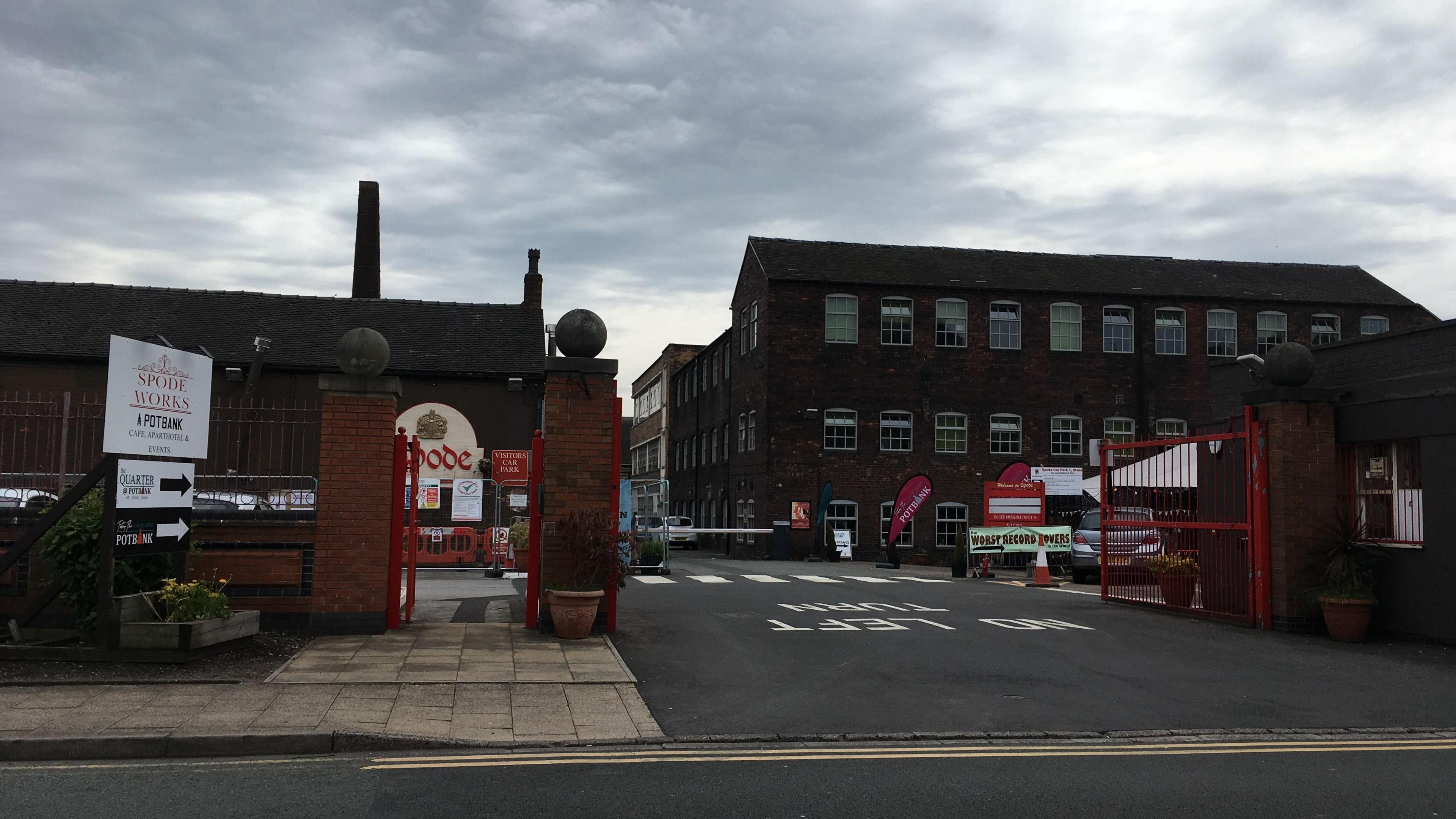 The outside of two brick buildings. One on the right has lots of small windows and sits behind an open red gate. On the left, the building has a large, long chimney and a white sign depicting a gold crown and the word "Spode". There is a white sign in front of it that reads "Spode works, a potbank"