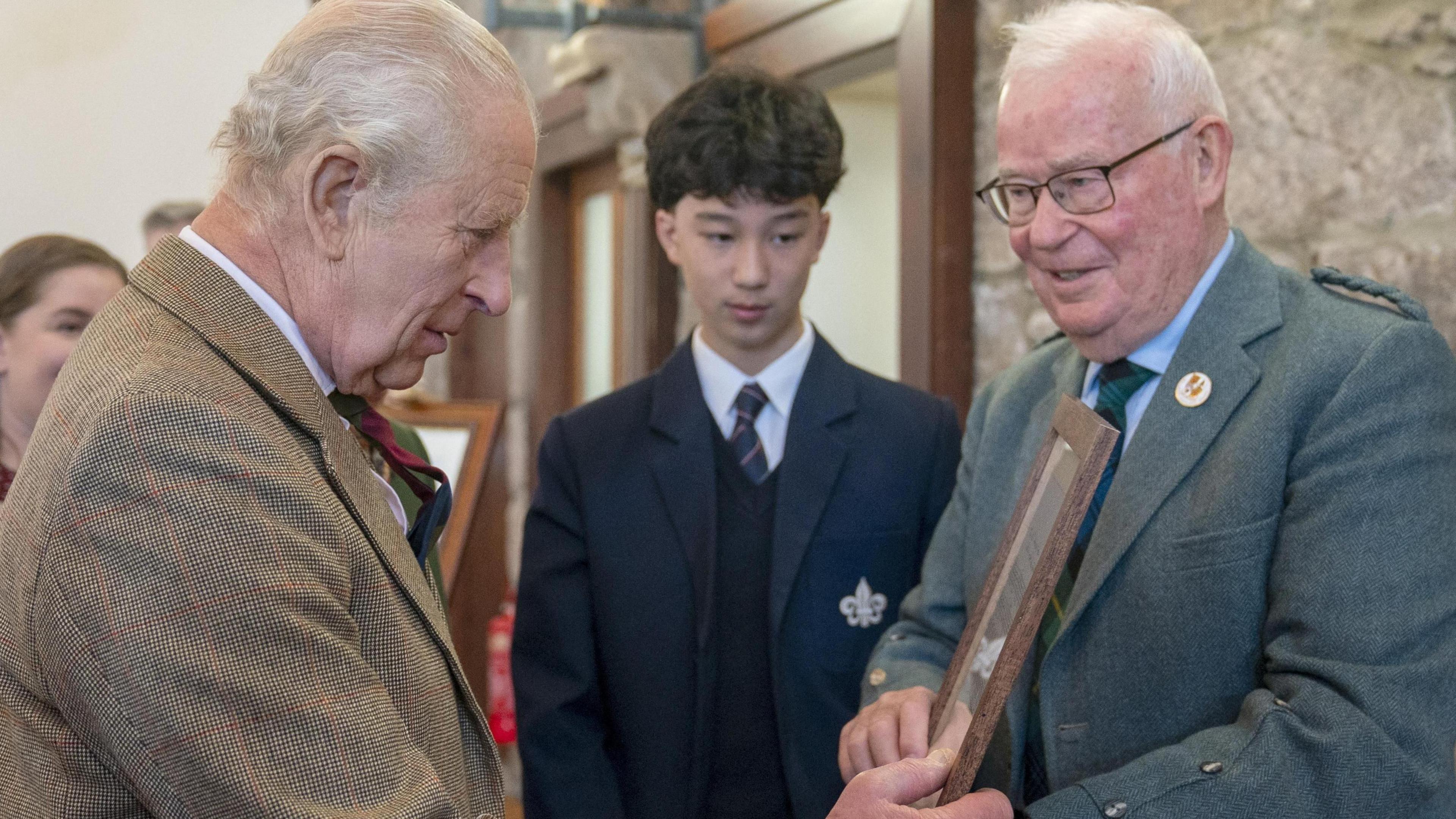 King Charles III, who has white hair and is wearing a brown tweed jacket, is presented with the framed score of a fiddle tune by Gordon Riddler, who also has white hair and is wearing a blue/green tweed jacket and a blue shirt with tartan tie.