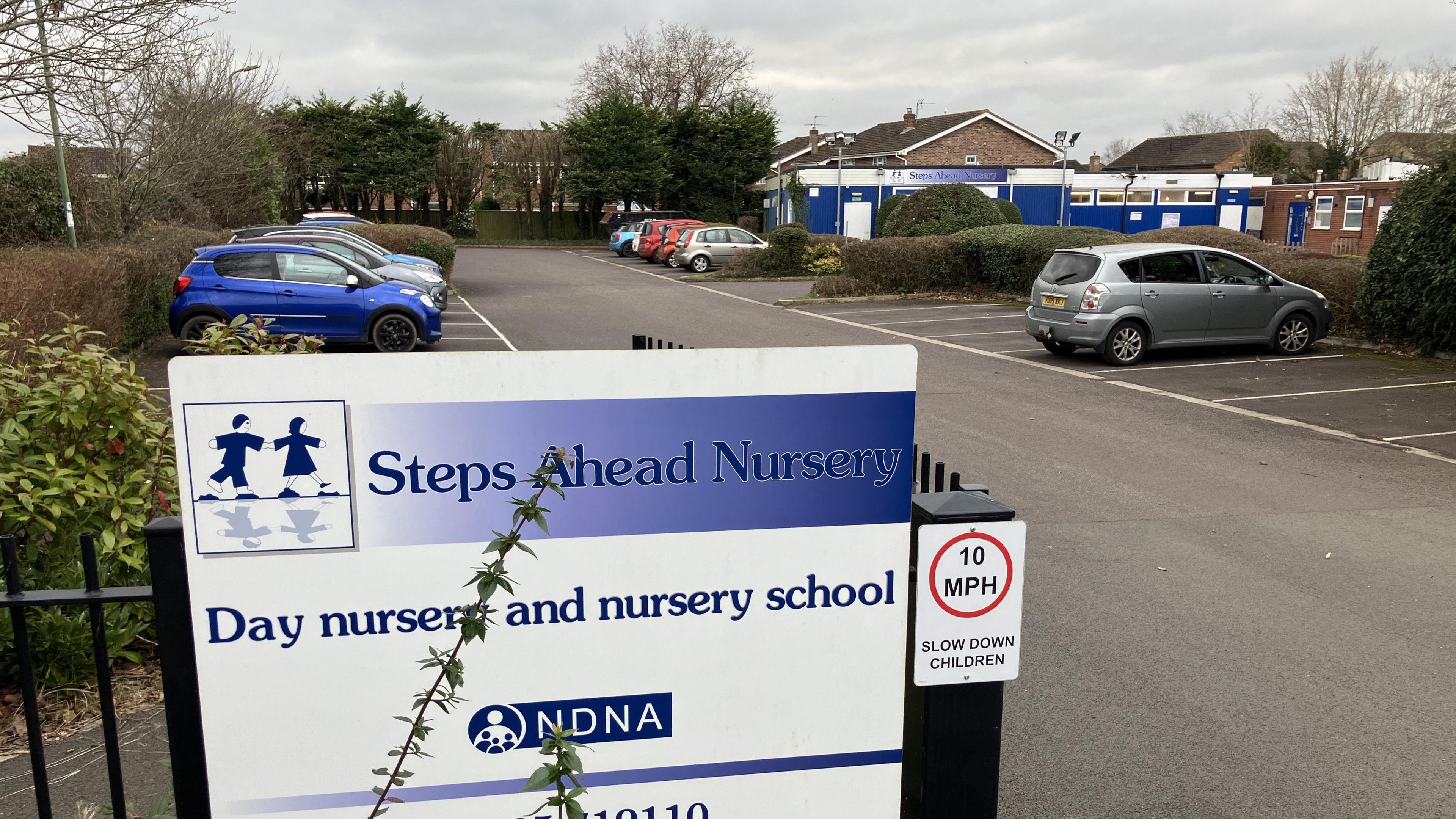 A blue and white sign for Steps Ahead Nursery, which is attached to a black fence on the perimeter of the car park. In the background you can see the nursery, which is a one-storey blue building with high windows.