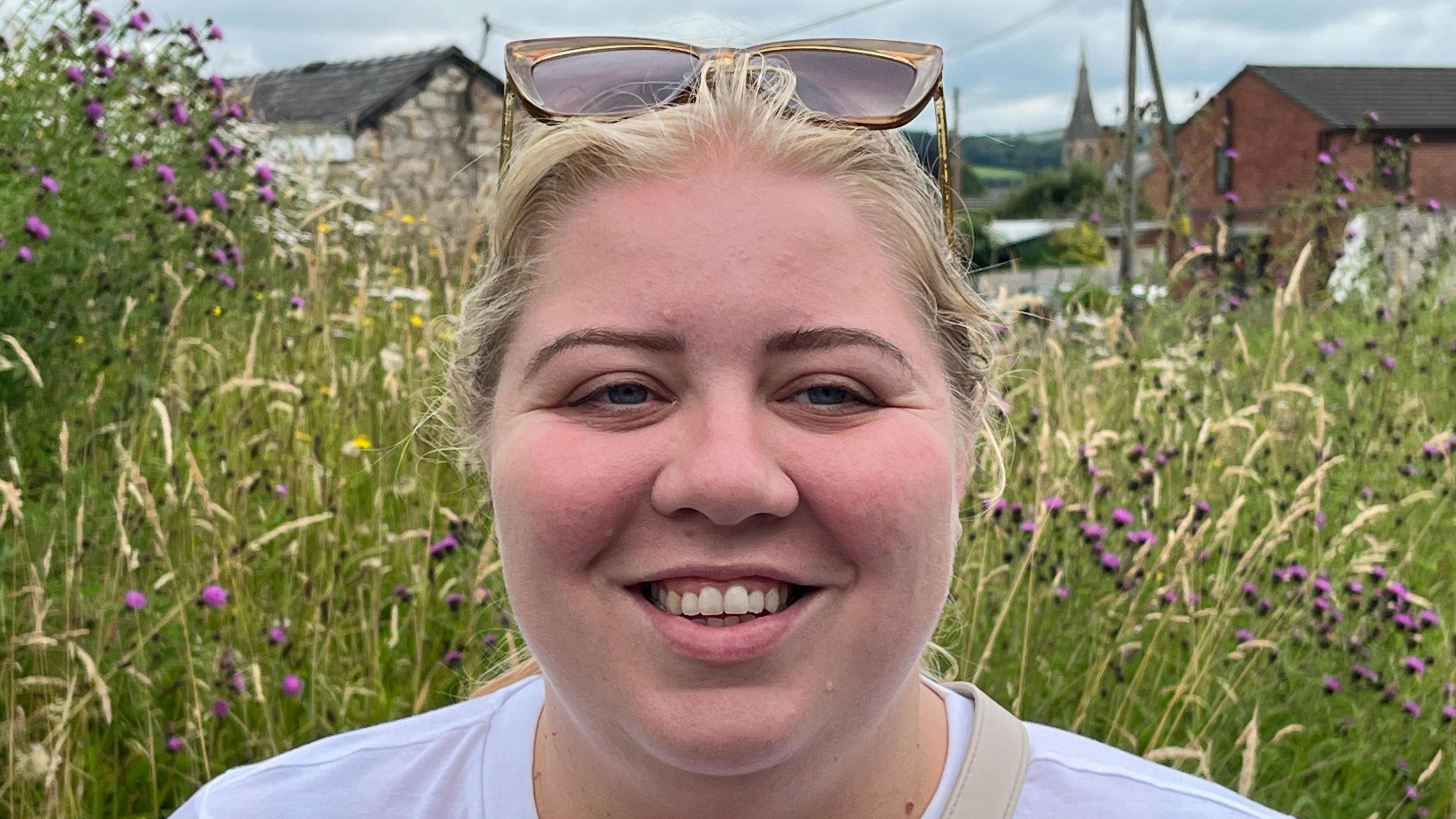 Emily Pierce with sunglasses on top of her head, wearing a white t shirt and standing in front of a field of grass and purple flowers with houses in background