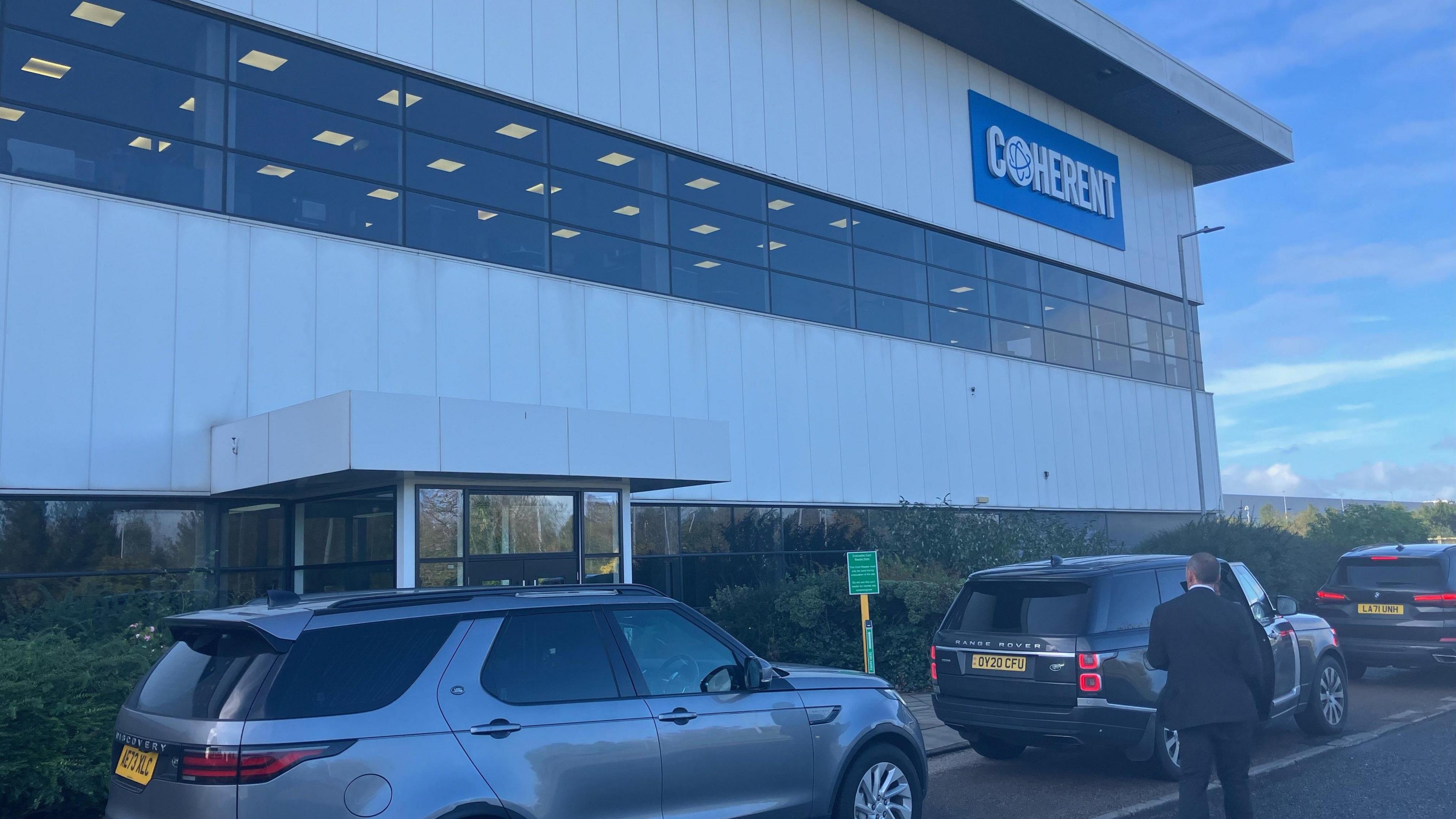 The semiconductor factory in Newton Aycliffe. It is a large, two-storey, modern industrial building with long, horizontal rows of dark glass windows between silver panels. Three cars are parked in front of it and a man in a dark suit is seen from behind.