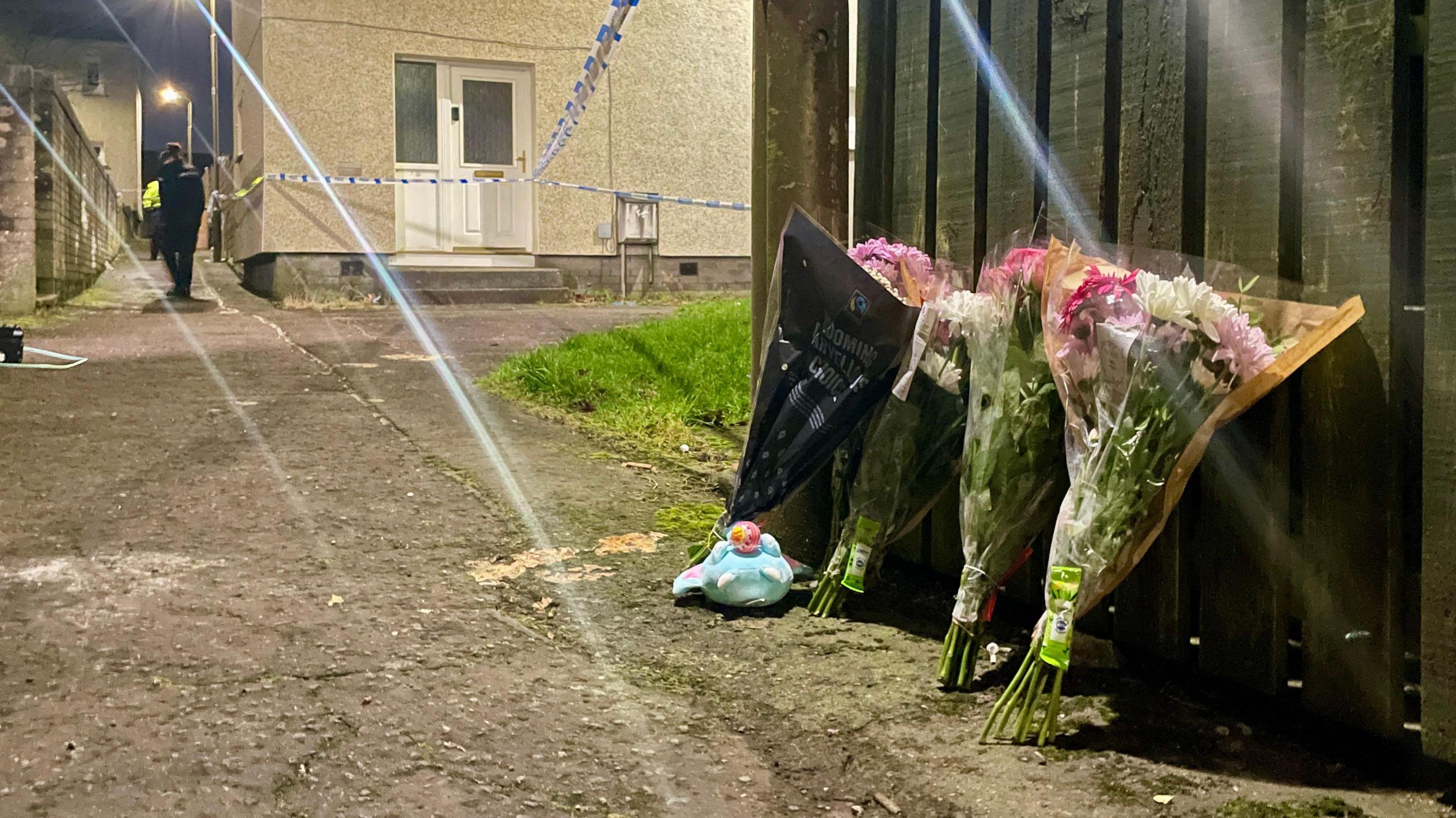 Floral tributes propped against a fence. Police officers can be seen in the background.