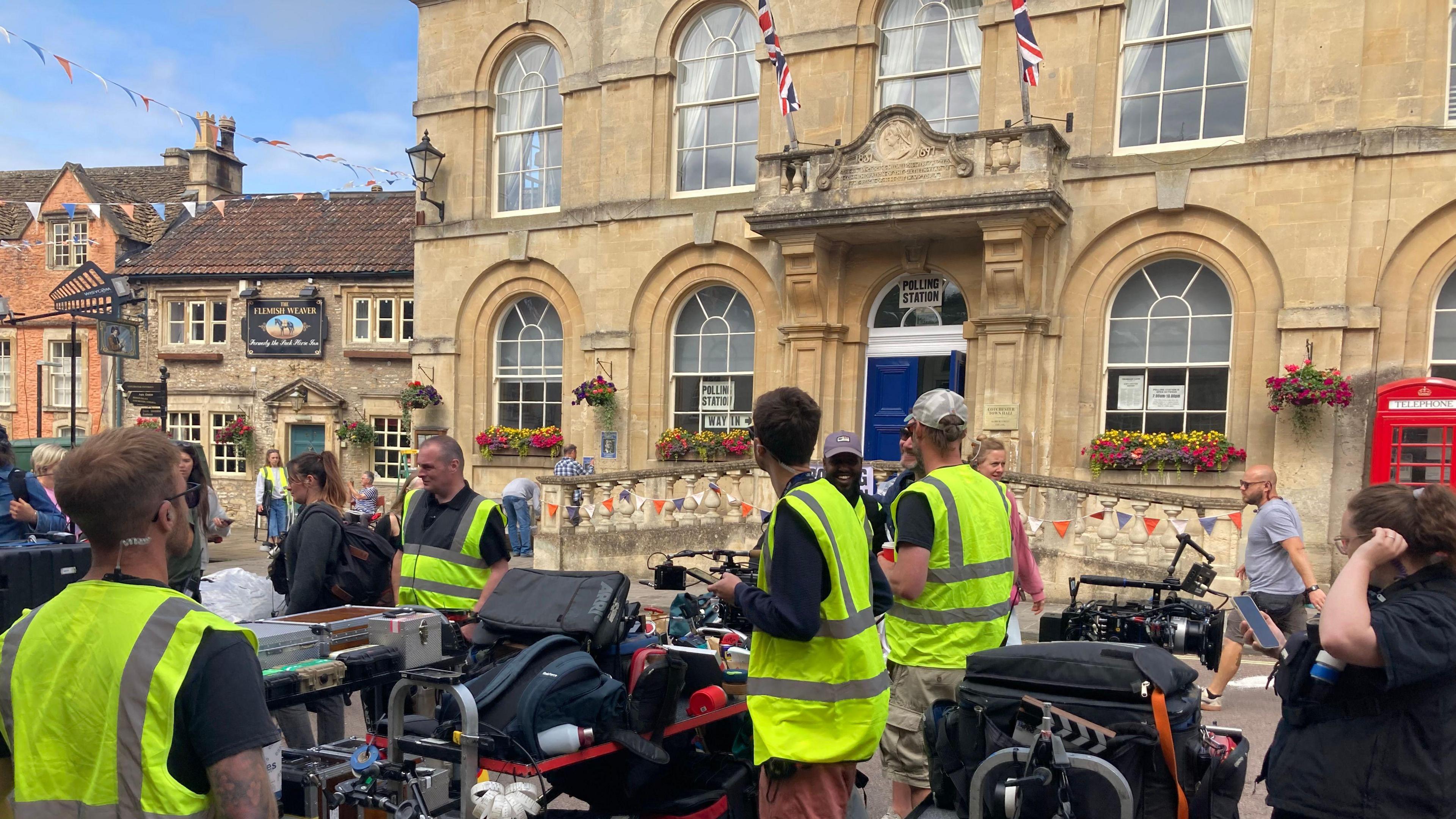 People in high-vis with camera equipment stand outside a grand-looking town hall made from Cotswold stone.
