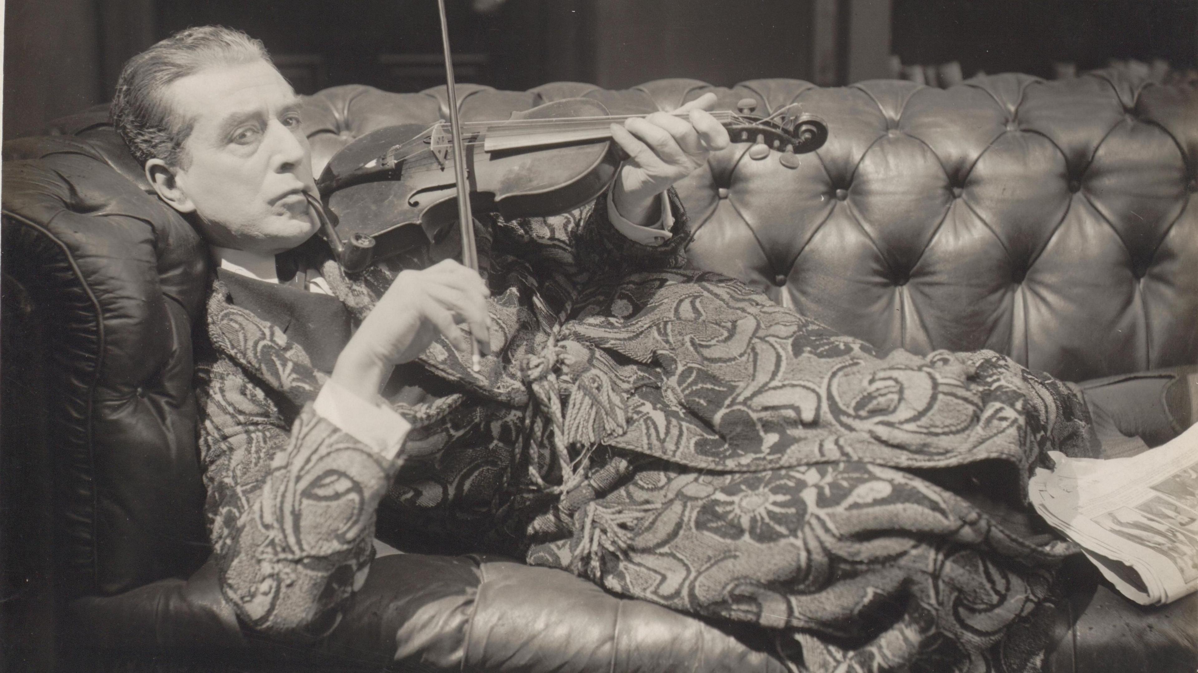 Eille Norwood, playing Sherlock Holmes, wearing an elaborately patterned dressing gown lounges on a leather couch, while playing a violin and smoking a pipe