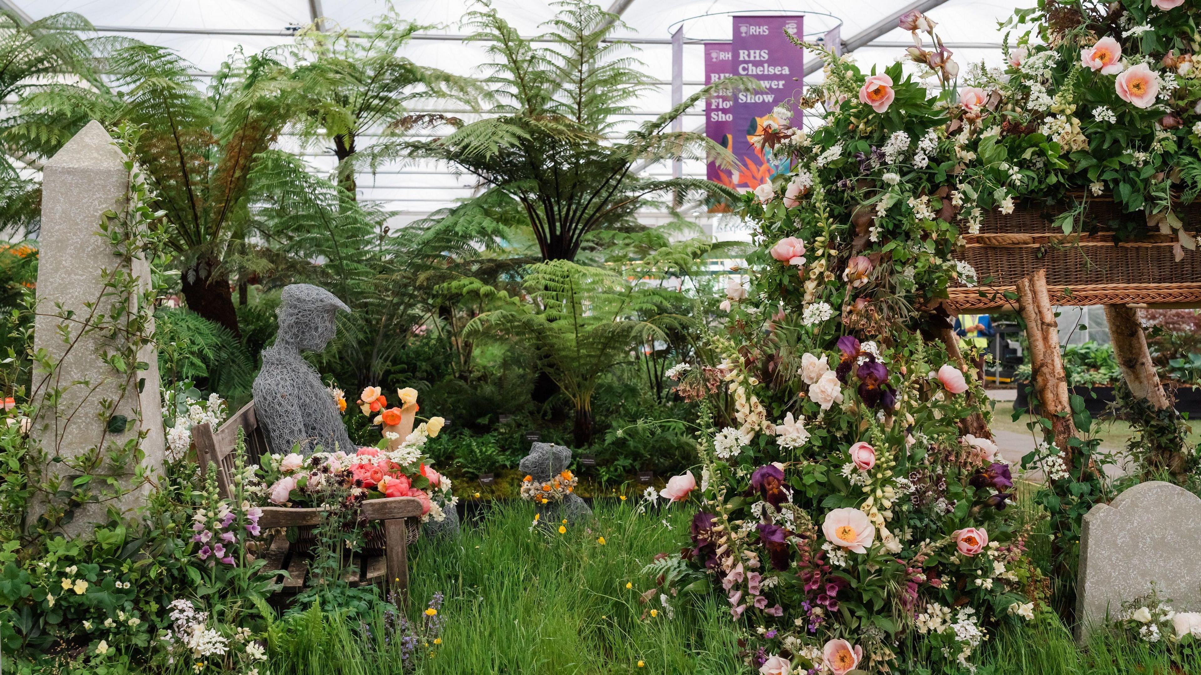 A flower display, with lots of brightly-coloured flowers flowing out of what looks like a wicker coffin near a gravestone. On a bench decorated with more flowers and surrounded by grass is a figure of a man wearing a flat cap, made out of wire, looking down at a wire dog sitting in the grass. The dog has flowers around its neck.