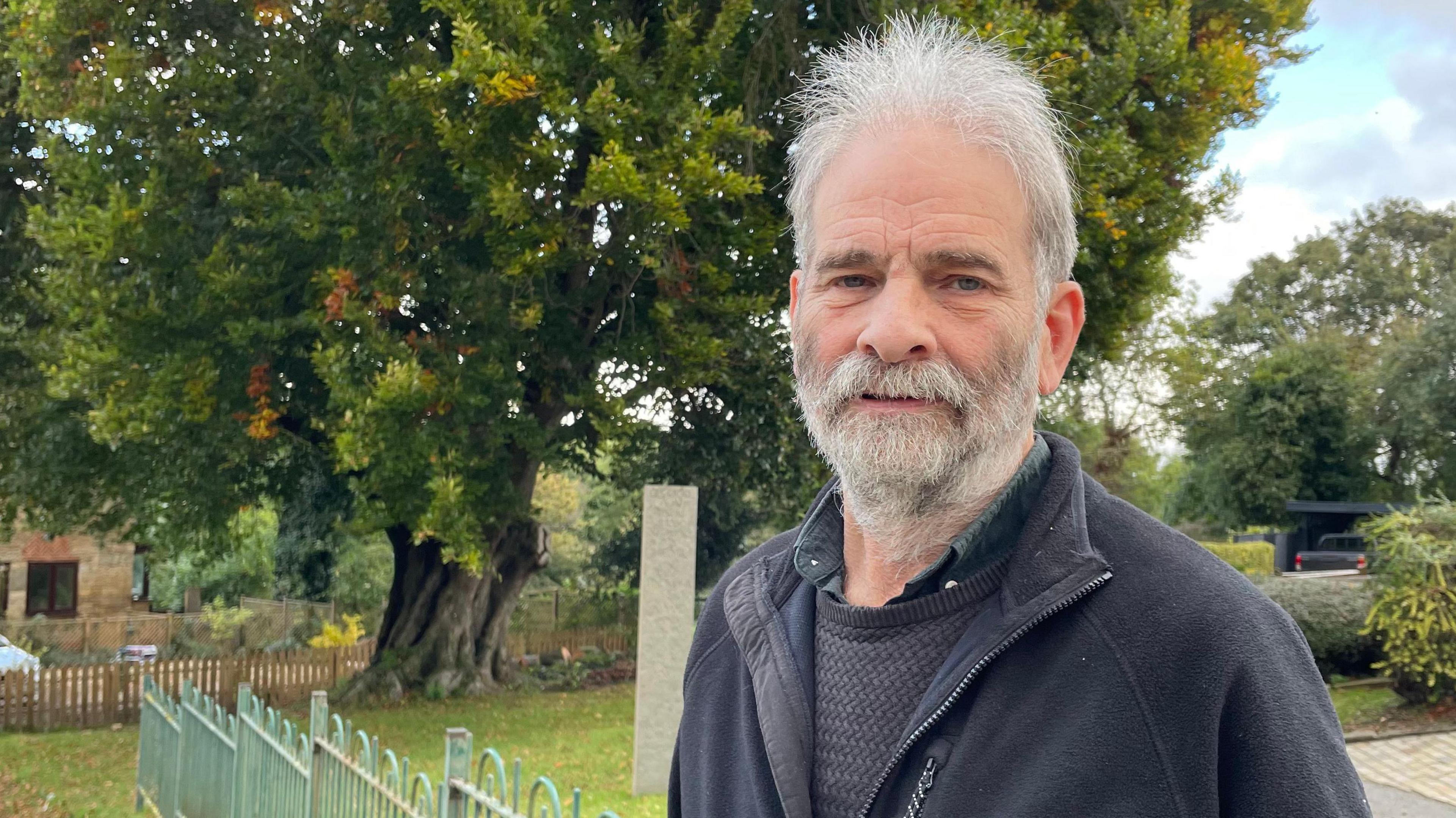 Nick Ballinger is stood in front of the beech tree. He's in his seventies and has grey hair with a beard.