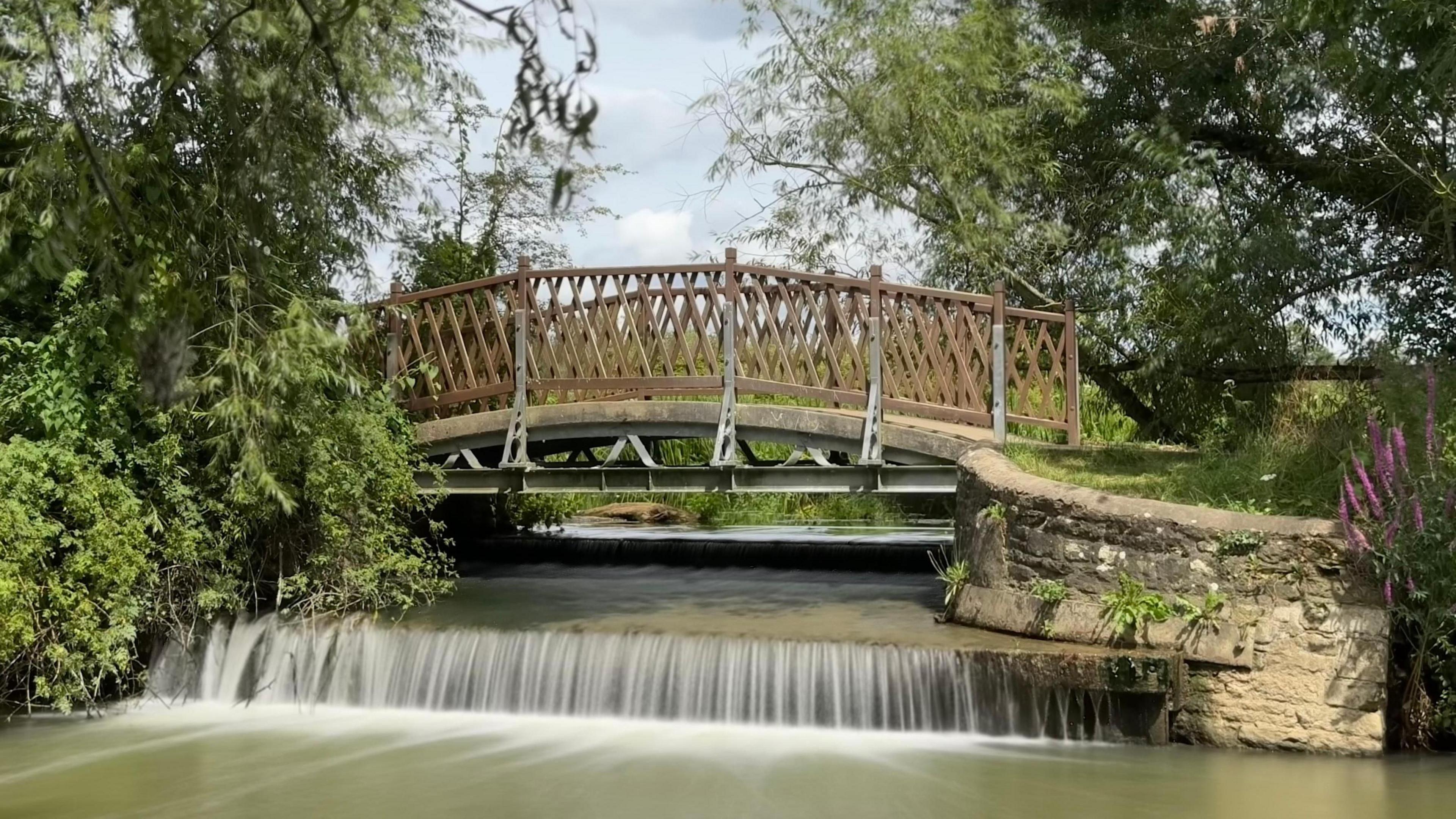 Water runs over a weir underneath a brown wooden bridge with a stone wall to the right and trees either side