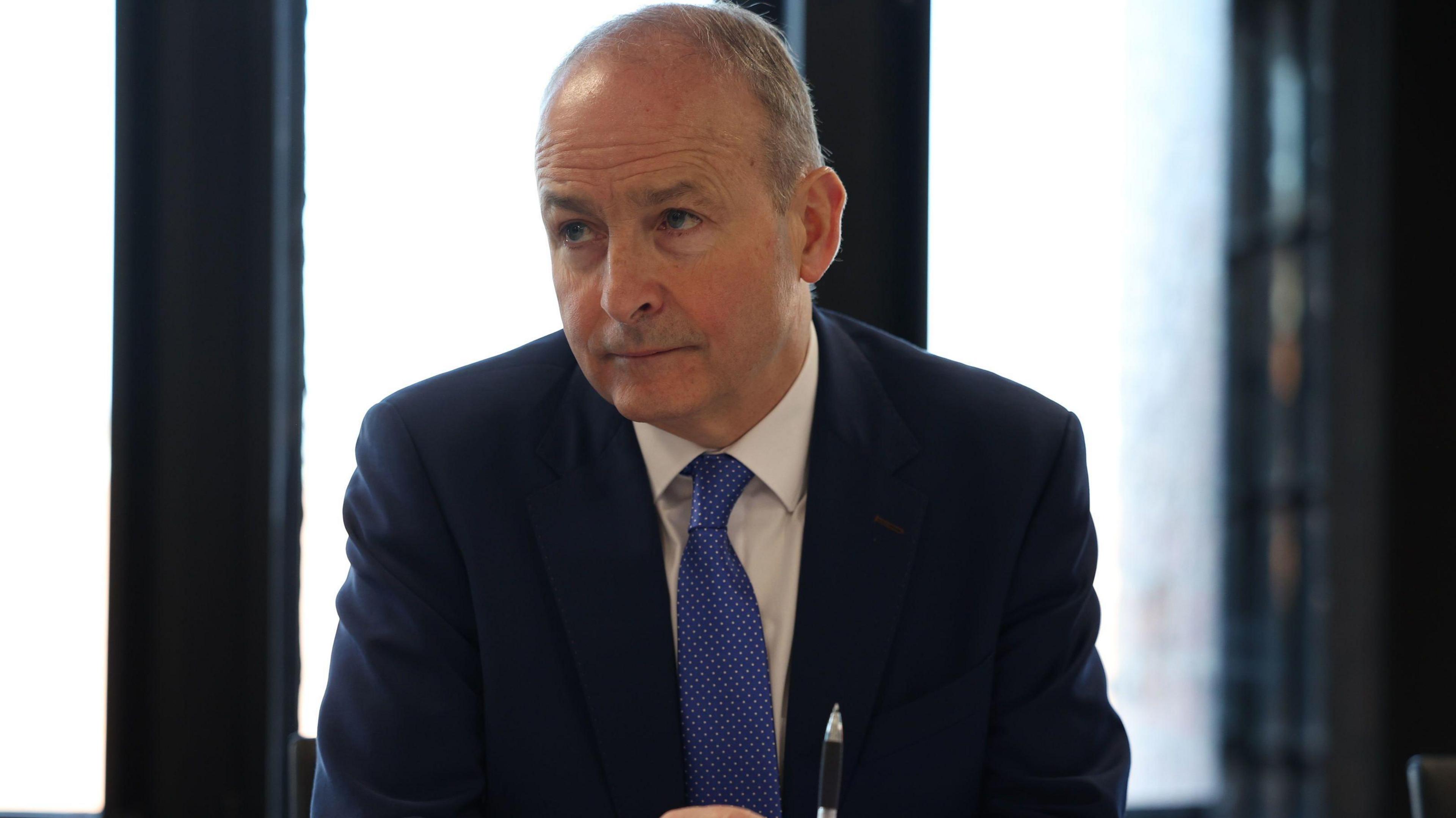 Taoiseach (Irish prime minister) Micheál Martin wears a blue suit, white shirt and blue tie. He has grey hair. He is sitting at a table.