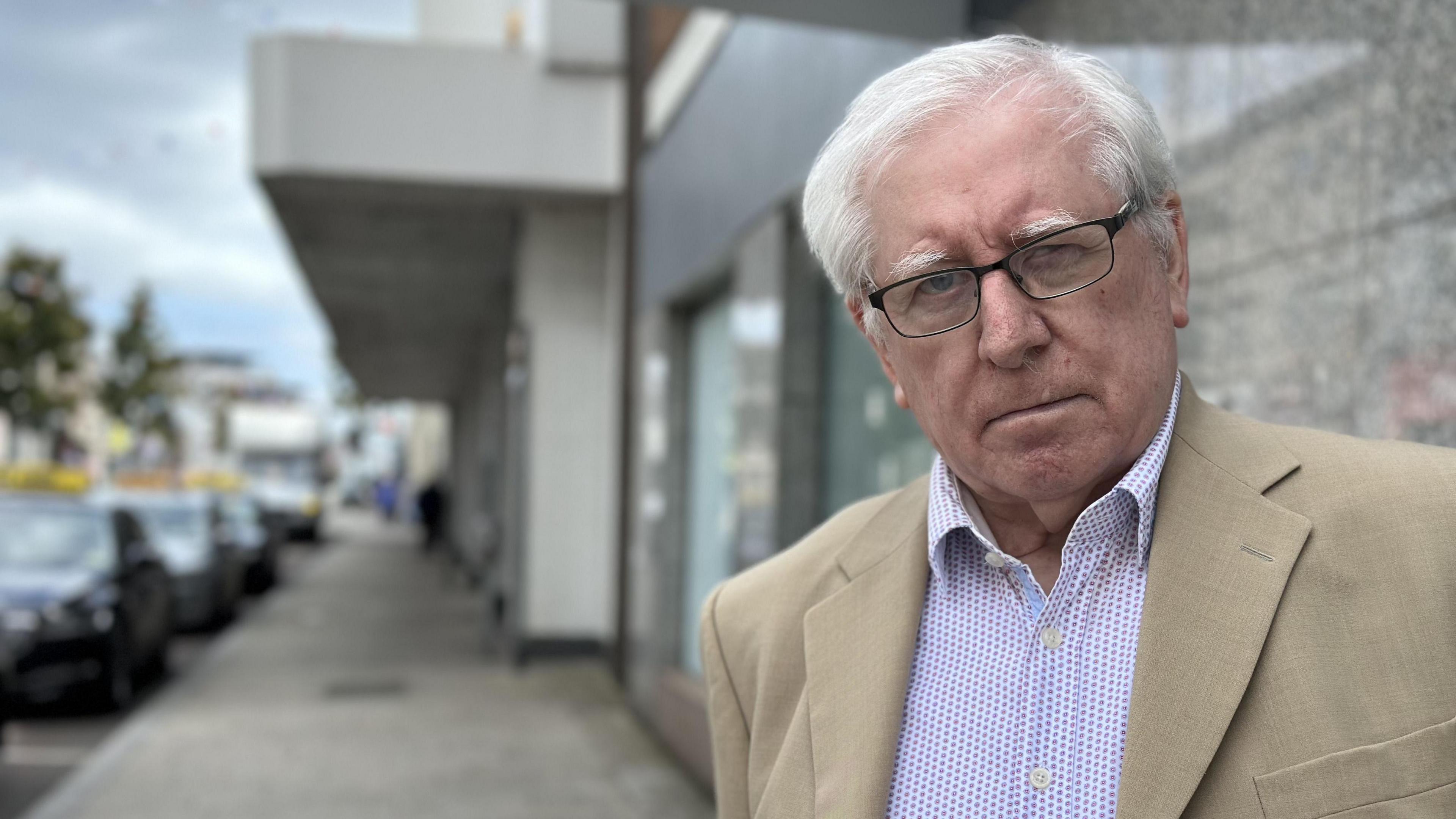 A white haired man pictured to the right hand side of the shot. He wears rimmed glasses, a beige suit jacket and a spotty shirt.