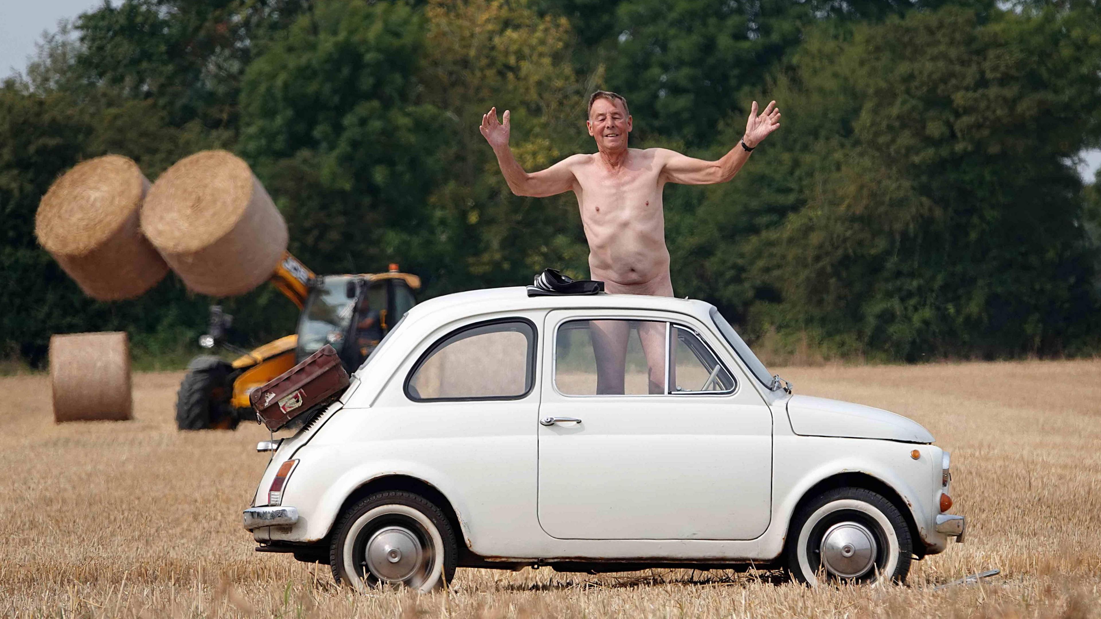 A naked man with his private parts obscured by a small white car. He is in a field with a hay baler in the background. 