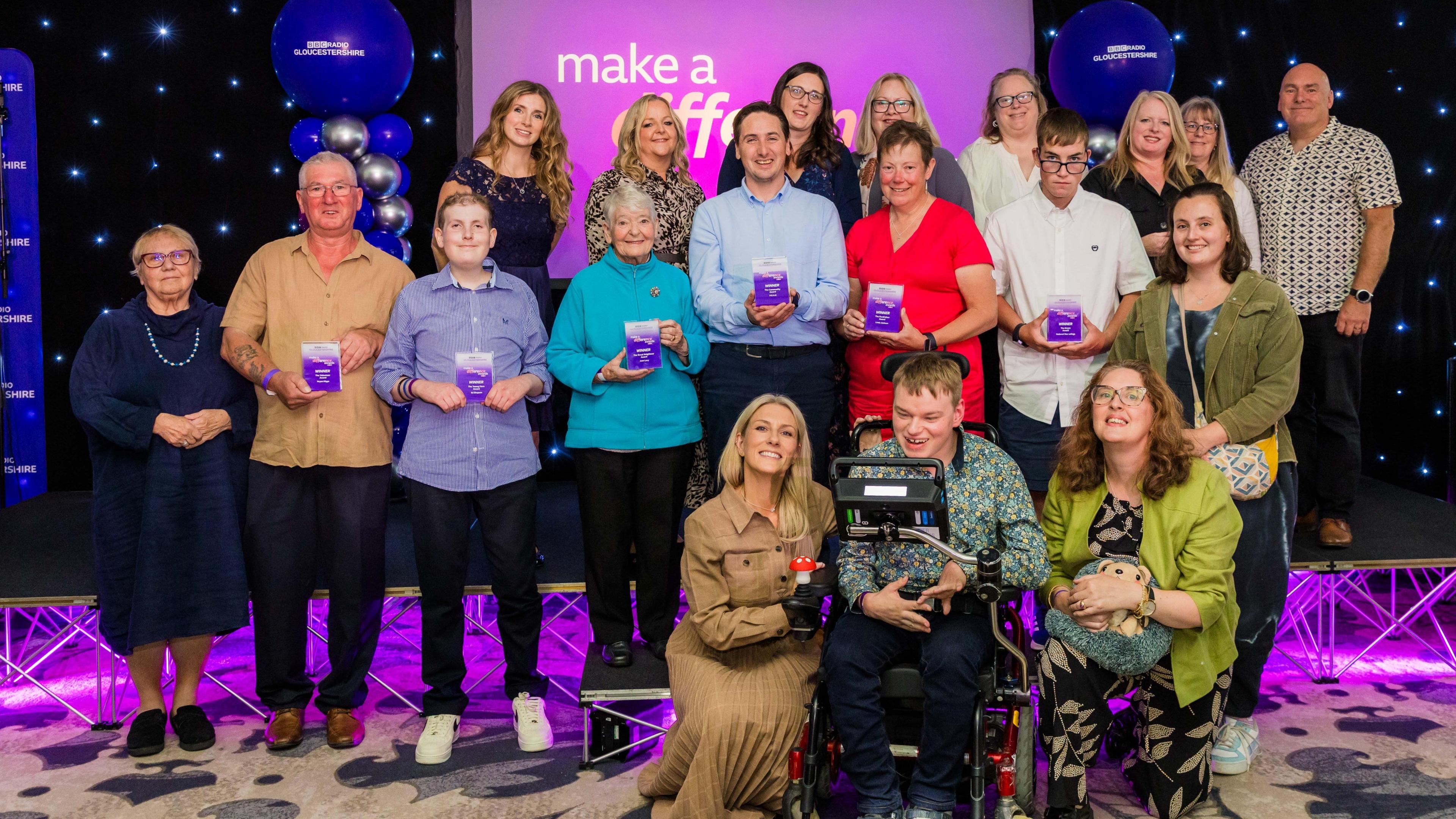 A group of about 20 award winners - a variety of people - stand holding their purple awards in a purple-themed conference room against a lightbox that says "Make A Difference"