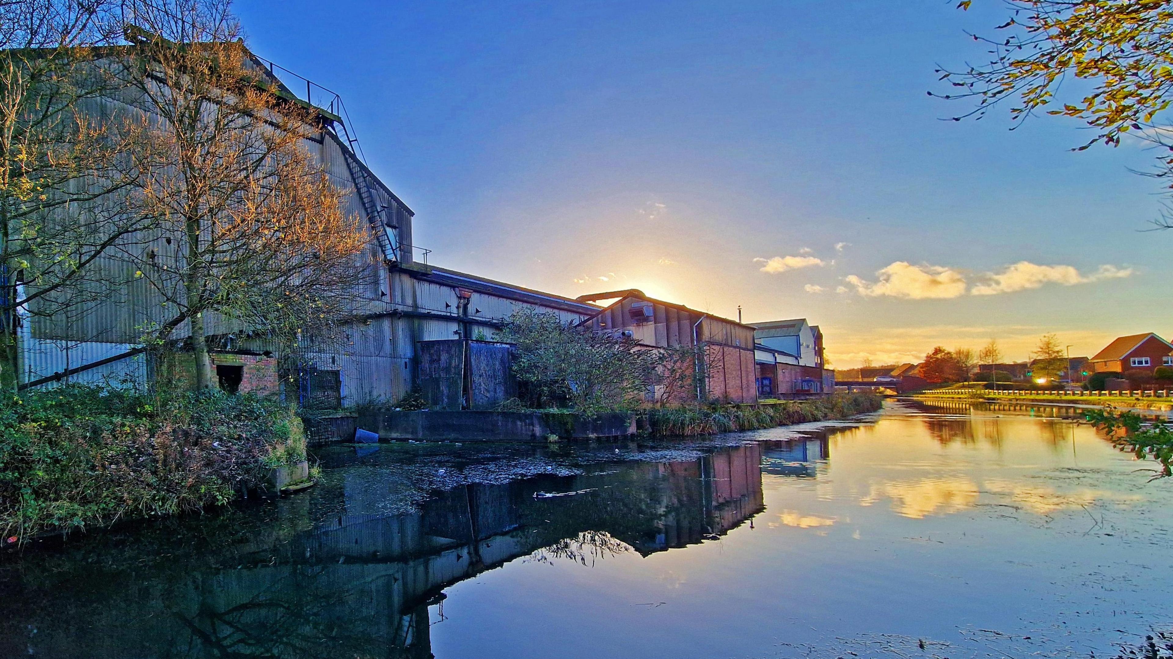 The sun is setting on a canal with clouds reflecting on the water. There are grey and red buildings sitting on the left side of the canal. 