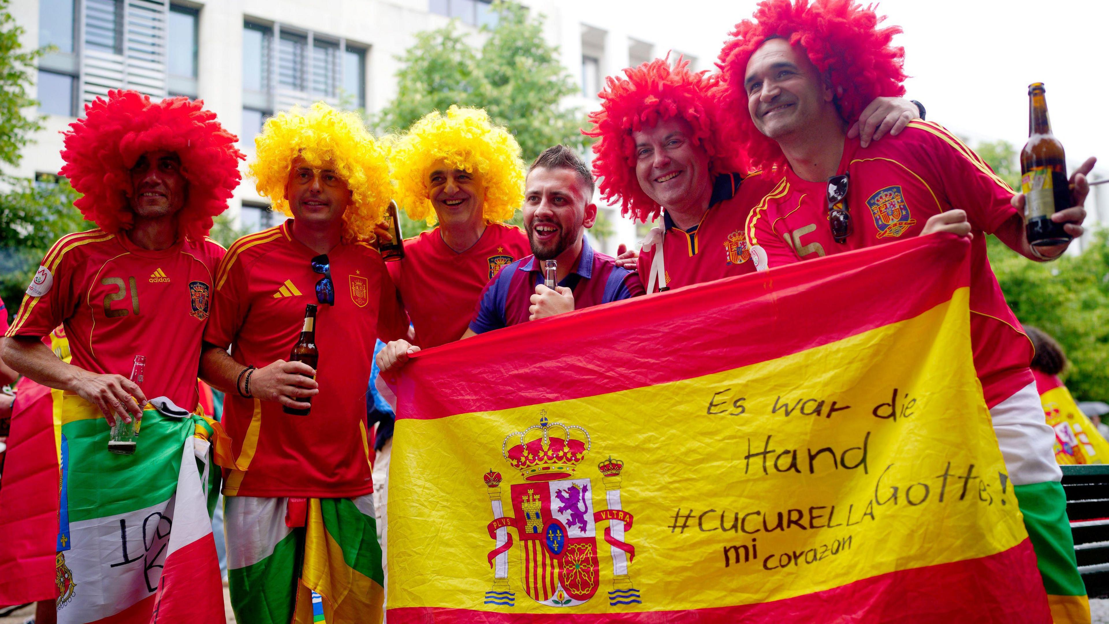 Spanish fans smile with a Spanish flag with bright red and yellow wigs