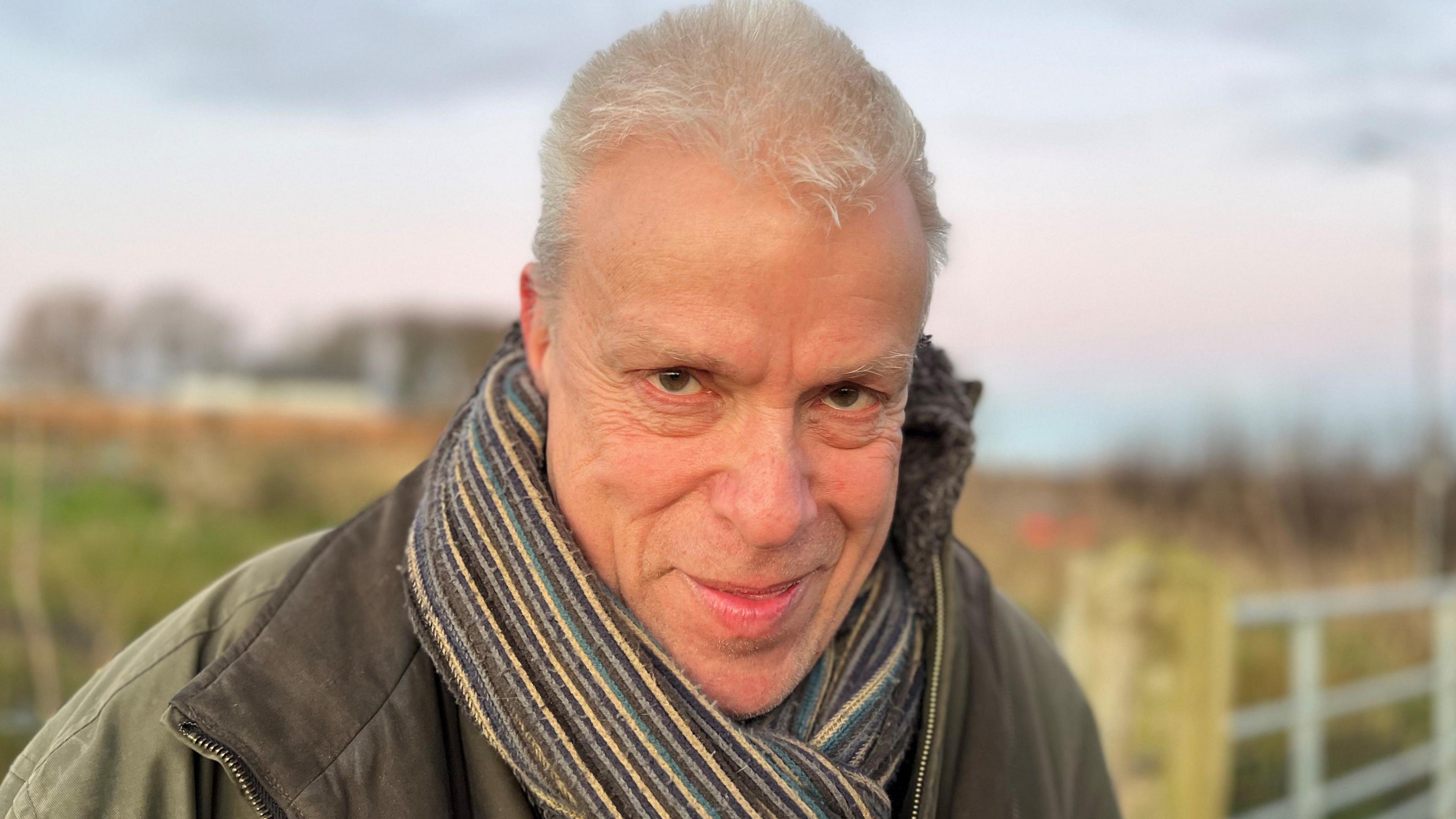 Nick Danby with grey hair and brown eyes smiling wearing a green coat and multi-coloured scarf as he stands in front of a fence to a green field