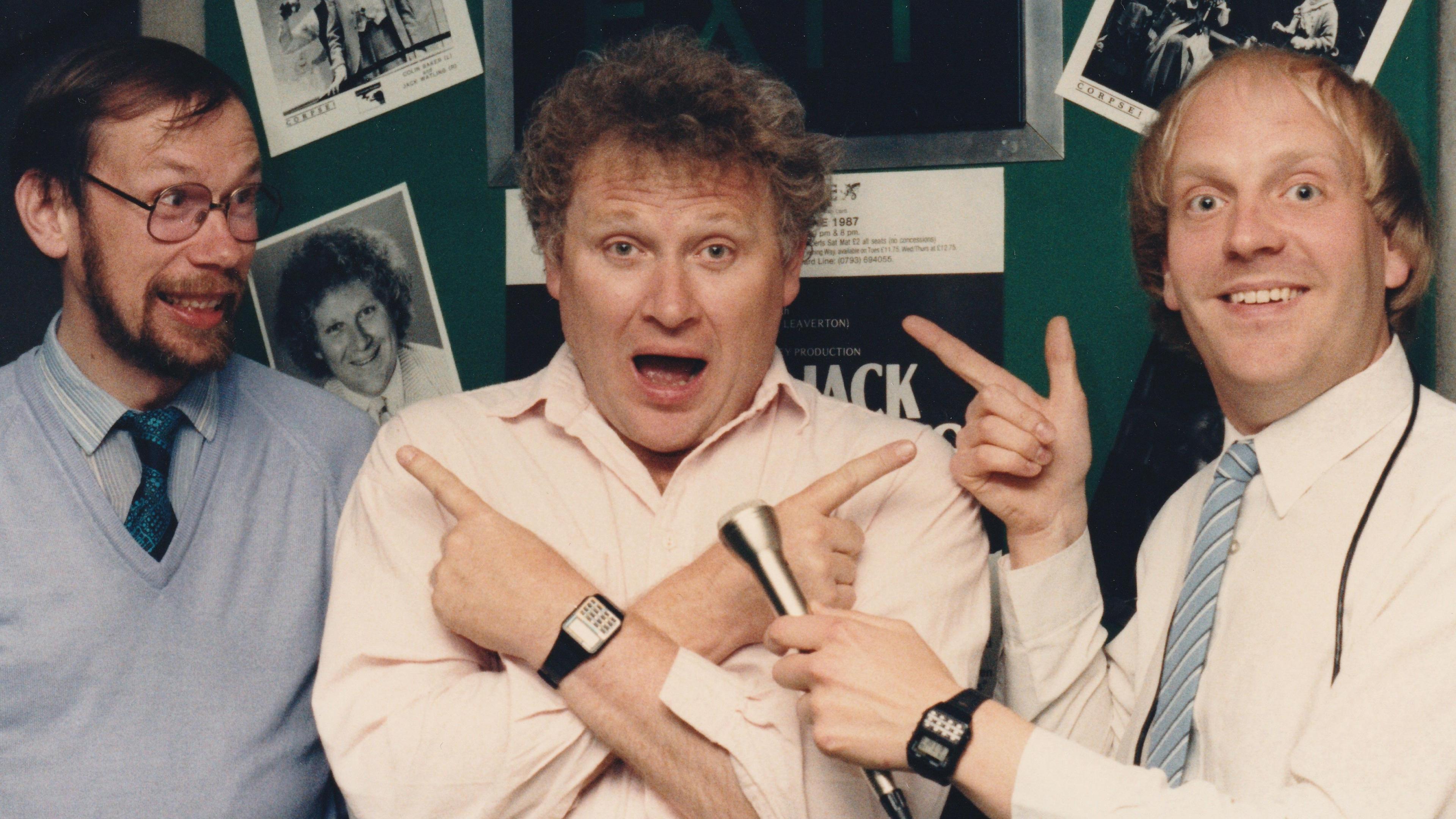 Archive photo of Dave Latham holding a microphone in front of Colin Baker and pointing at him. Another man is on the other side of Mr Baker