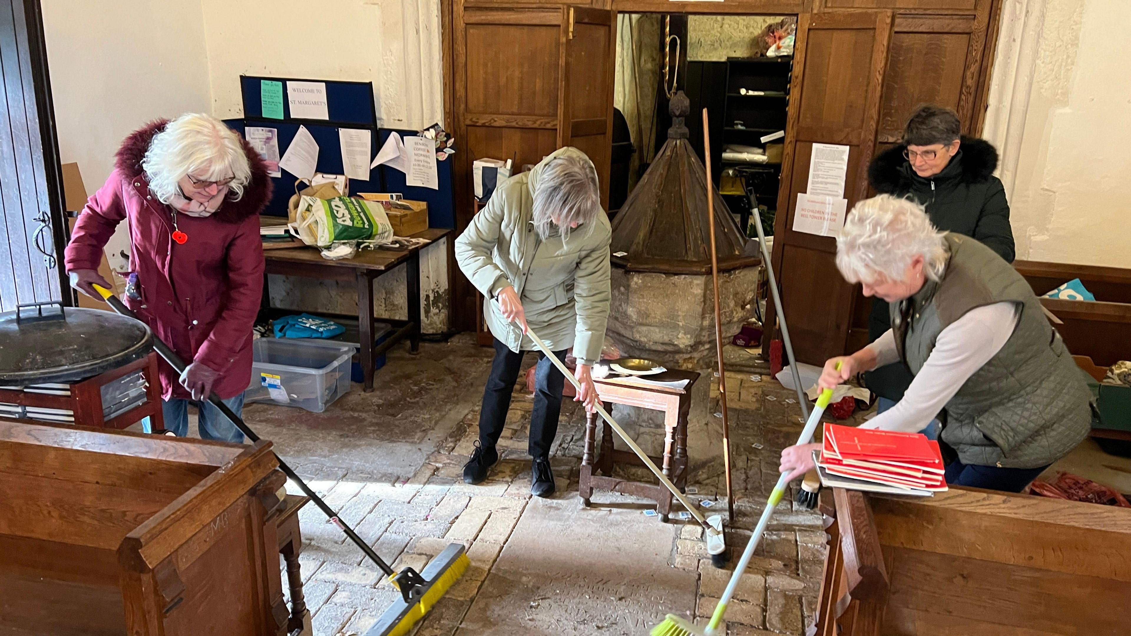 Three elderly woman with brooms sweeping the interior church floor and between pews.