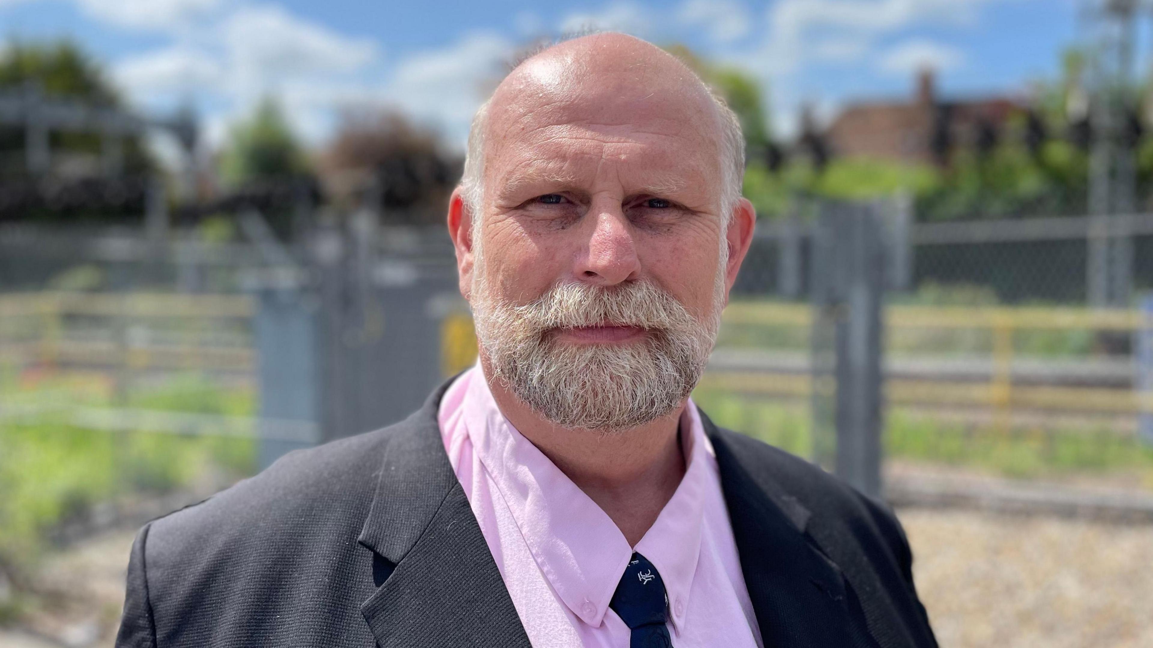 Tom Wootton, with very short grey hair and beard. He is wearing a dark grey jacket, pink shirt and dark tie and is looking at the camera. He is standing in front of a grey metal fence with grass and hedges beyond.
