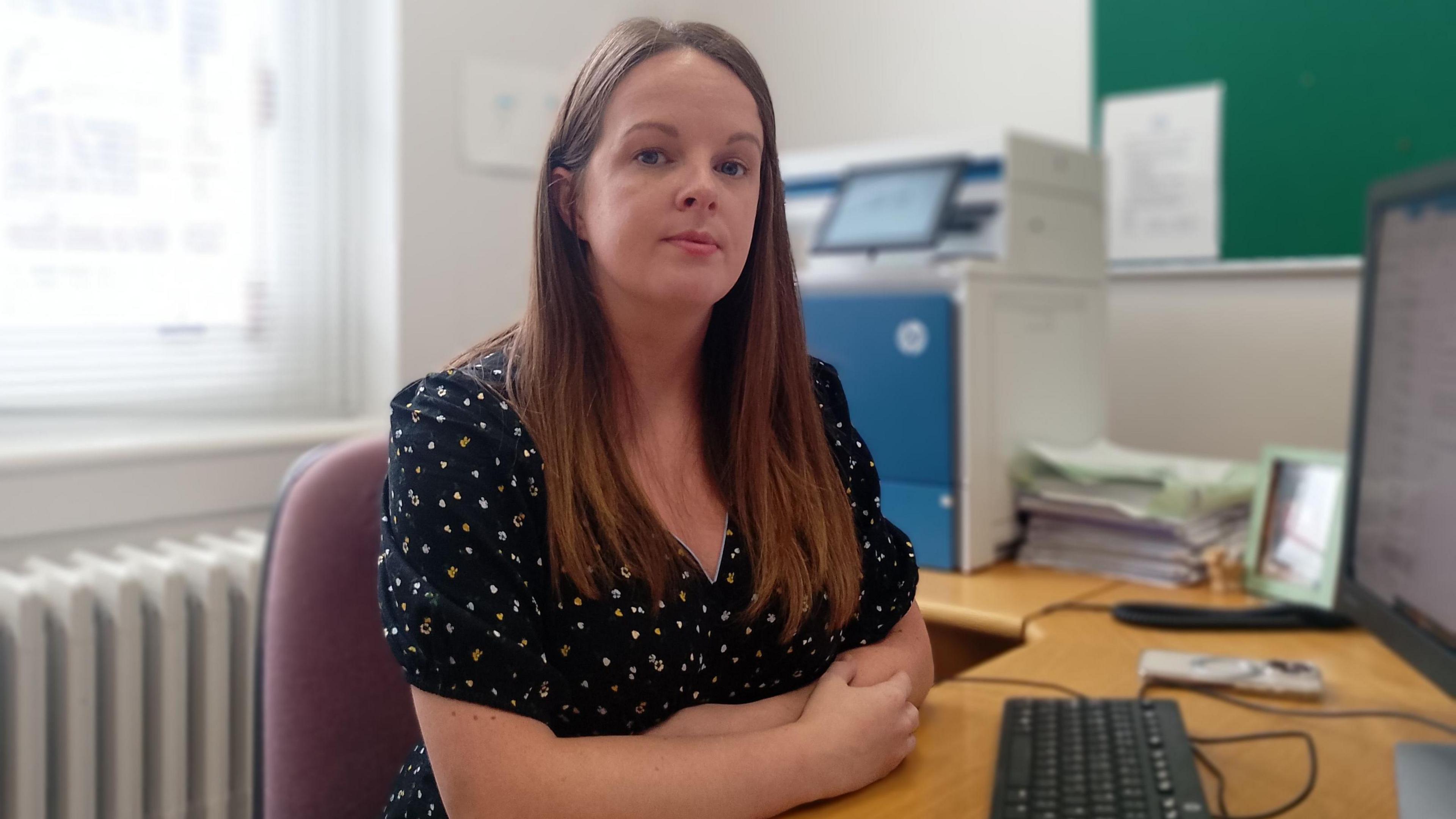  Nuala Mc Allister is sitting at a desk with a computer and keyboard in front of her. Her arms are folded. She has long brown hair. Paper, a mobile phone, a printer and a frame are sitting on the desk.