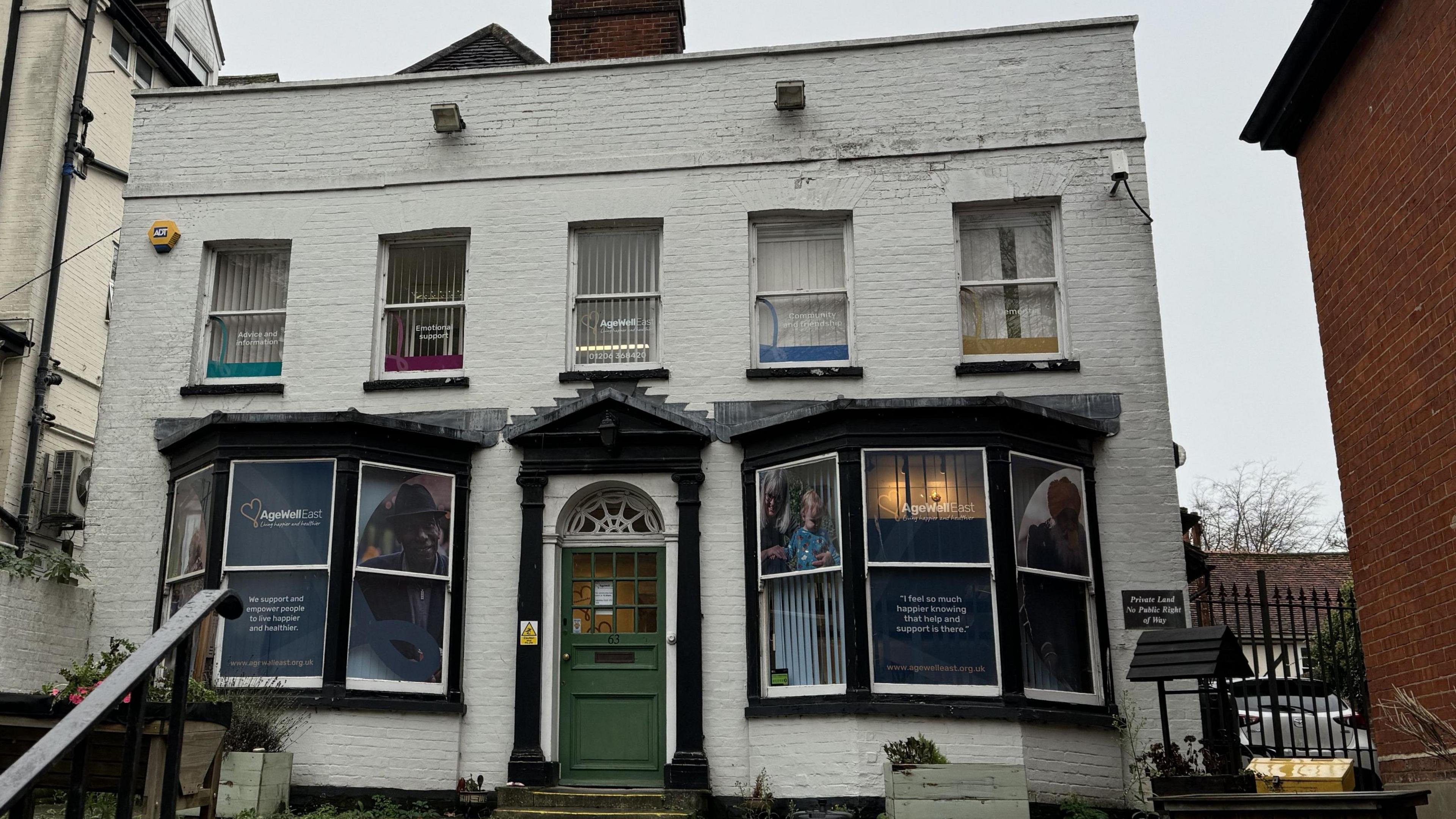 A white brick building with a green door. Age Well East signs can be seen within the windows