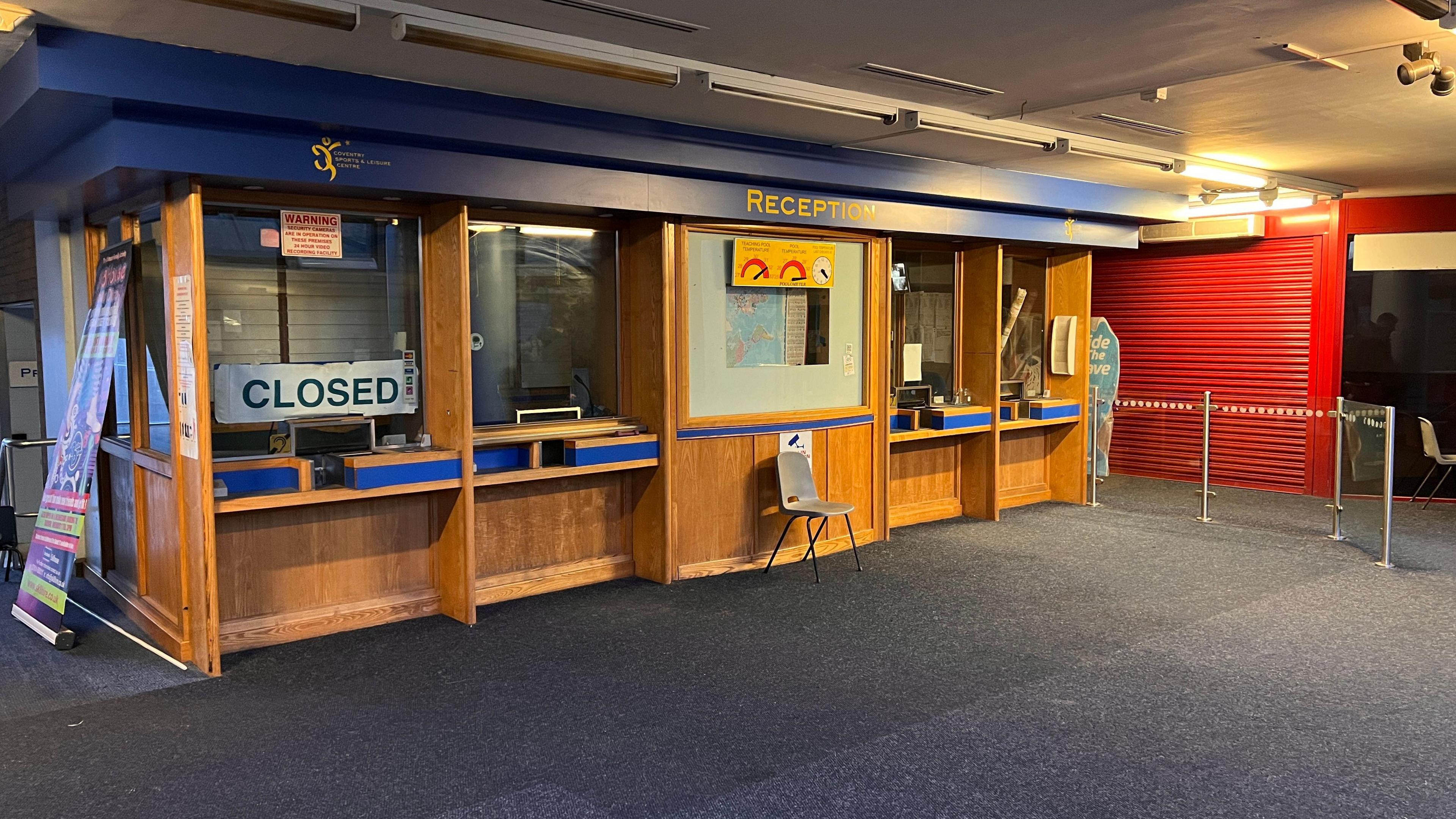 The abandoned reception area at Coventry Sports and Leisure Centre. The signage is blue with white writing. Four serving windows are visible, the one on the far left displayed a large closed sign in capital letters in blue writing on a white background. There is grey carpet leading to the area and the serving windows are surrounded by a light wood structure. Closed red shutters are visible to the right of the reception area.