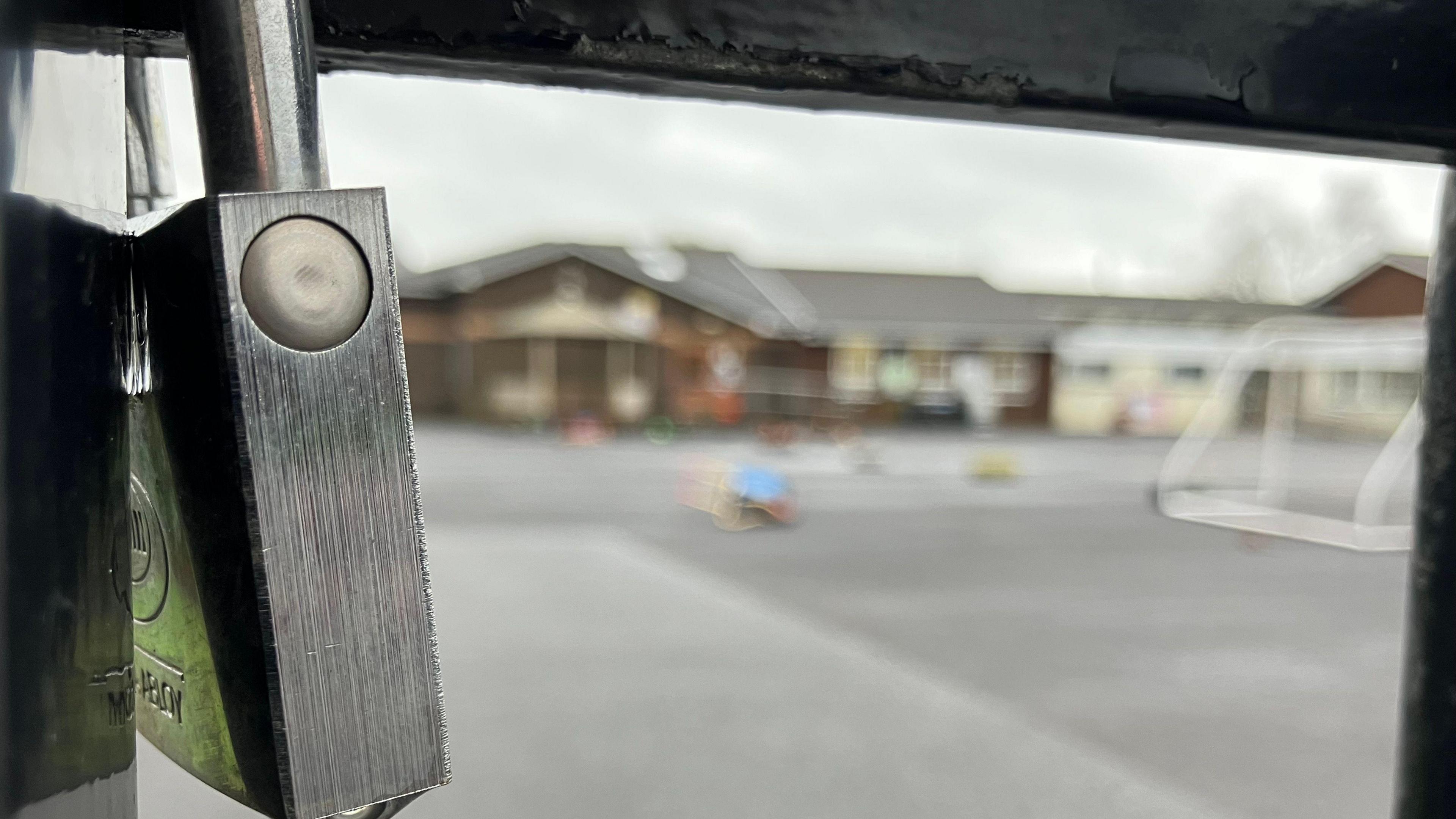 An extreme close up of a silver padlock attached to black gates. A school is out of focus in the background.