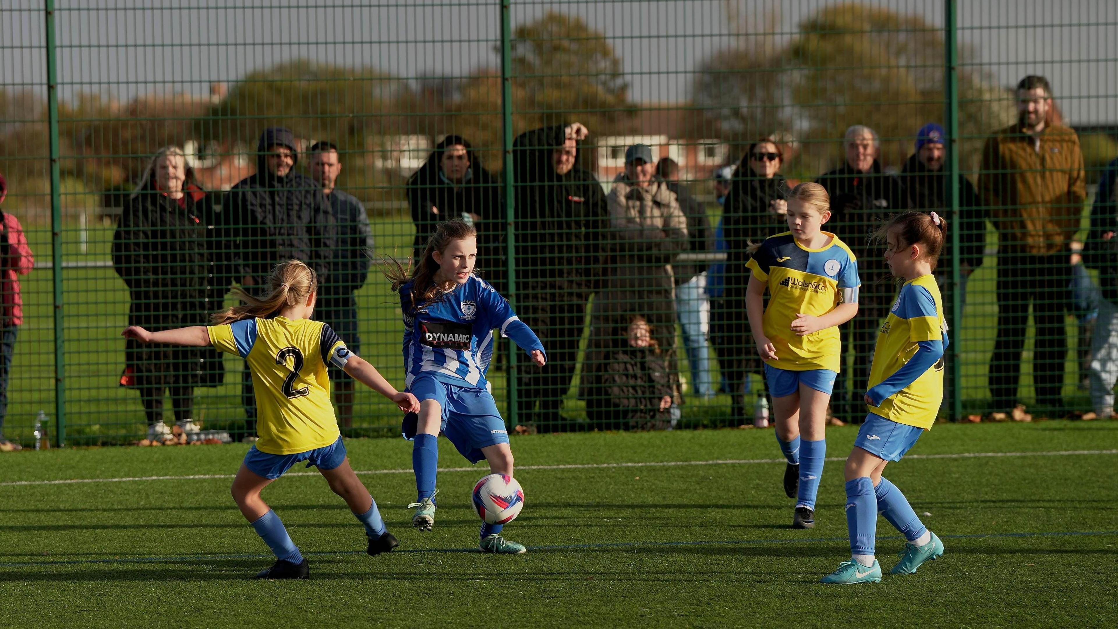 Four girls wearing the colours of Whitley Bay Football Club and Whitley Bay Sporting Club play on an artificial pitch with supporters standing behind a green metal fence.